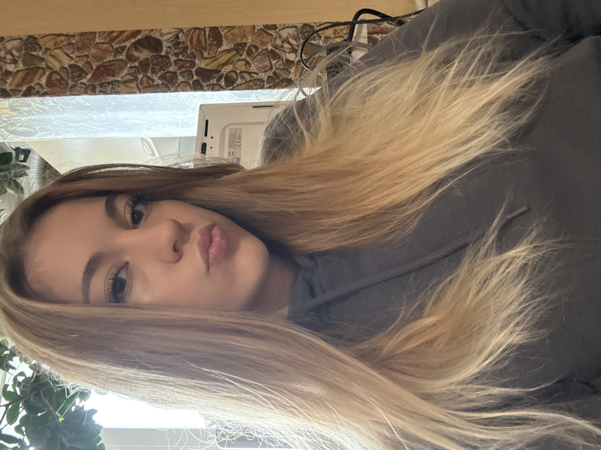 A young woman with blonde hair and fair skin is facing the camera with her lips pursed. She is wearing a gray hooded sweatshirt. Behind her is a wall with a stone pattern and a window with a plant in front of it. A white electronic device with wires attached is visible near the top of the frame. The image is rotated. The location in the city of Sofia, Bulgaria is unknown.