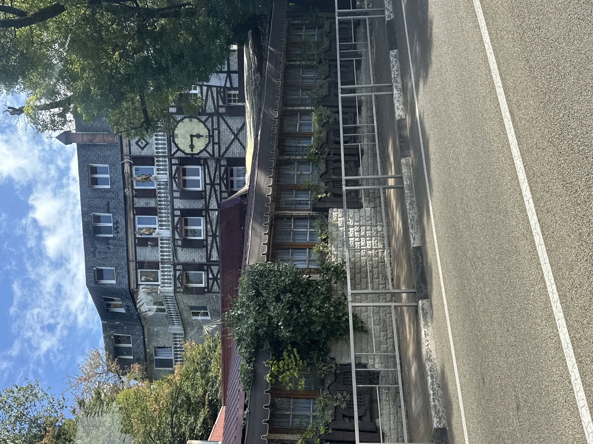 The image shows a multi-story building with a distinctive clock on its facade, situated in Sochi, Russia. The building has a mix of stone and timber-framed architectural elements, with numerous windows and balconies. A covered outdoor seating area with awnings is visible at the ground level, featuring dark wooden seating and potted plants. A metal fence separates the building's grounds from a paved road in the foreground. Green trees and foliage surround the structure, suggesting an outdoor setting. The sky above is blue with scattered clouds.