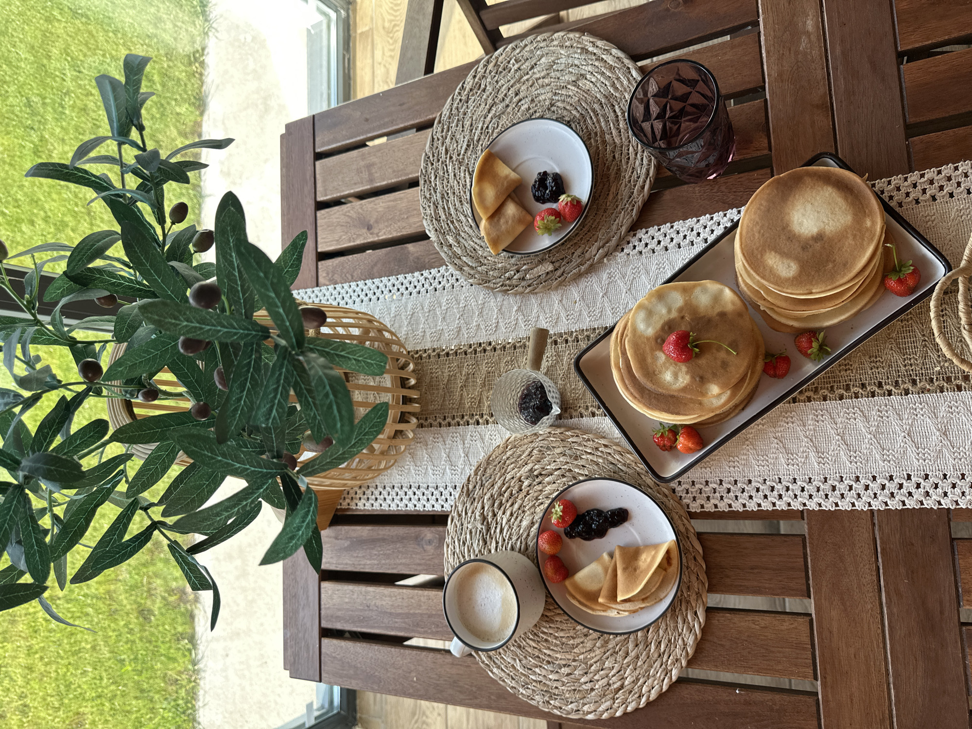 A high-angle medium shot captures a breakfast setting on a wooden outdoor table, likely on a balcony or patio in the city of Samarsky, Russia. The table is set with a light-colored woven placemat and a decorative white table runner. In the foreground, a stack of golden-brown pancakes is presented on a rectangular white plate with a dark rim. Several fresh strawberries are scattered around the pancakes, with one strawberry placed on top of the uppermost pancake. To the left, another plate holds a smaller stack of folded pancakes, three strawberries, and a dollop of dark jam. A white mug with a frothy beverage, possibly coffee or latte, sits on a round woven placemat to the left of this plate. A small glass bowl containing more dark jam is positioned between the two plates. To the right, a faceted dark pink glass contains a dark liquid, possibly juice or wine. In the background, a large artificial olive tree in a woven basket is placed centrally on the table, its leaves and dark faux olives obscuring a portion of the view. Beyond the table, a blurred background shows a patch of green grass and a window or glass door. The overall scene evokes a sense of a peaceful morning meal.