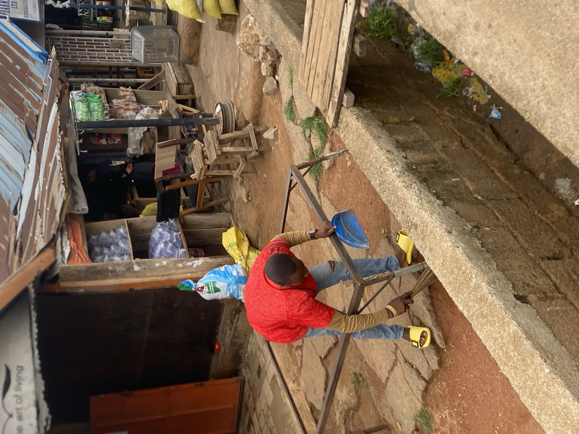 A man wearing a red shirt, blue jeans, and yellow sandals is cleaning debris from a concrete drain next to a street in Bukuru, Nigeria. He is using a broom and holding a blue plastic dustpan. The drain is filled with trash and some sparse vegetation. The street scene includes market stalls with various goods on display, including bags of grain, food items, and other merchandise. The buildings lining the street are constructed of corrugated iron and wood. The setting suggests a busy, informal market environment.