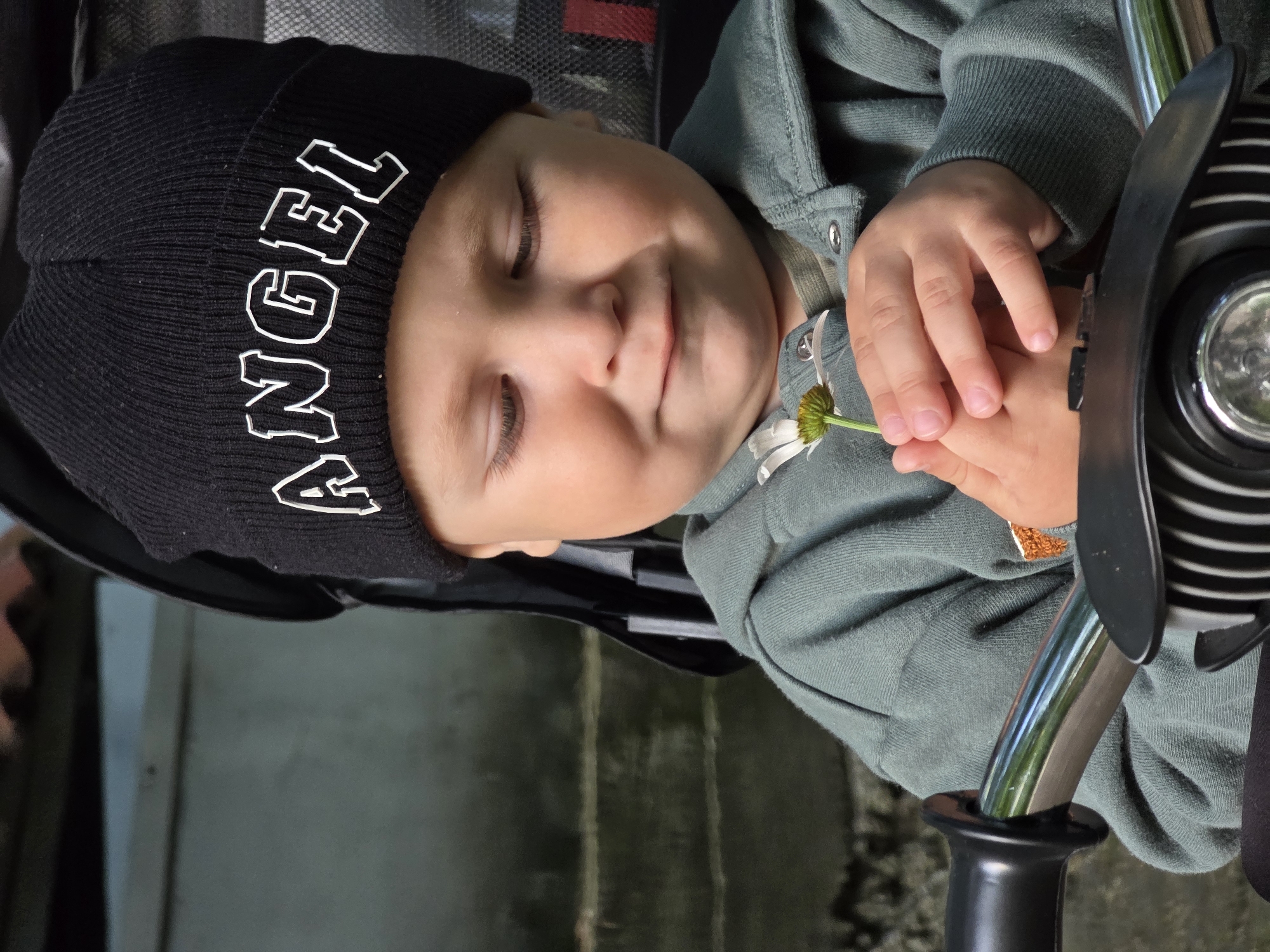 A close-up, eye-level shot shows a young child wearing a black beanie with the word "ANGEL" in white lettering, and a teal hooded jacket. The child is holding a small white daisy with a yellow center in their hands, looking down at it with a gentle smile. The child is positioned in what appears to be a stroller or a similar seating apparatus. The background shows an out-of-focus outdoor setting with textured grey steps and a dark roofline. The lighting is natural, creating soft shadows. The image appears to have been taken in an urban or suburban environment, possibly a park or a residential area in Chaikovsky, Russia.