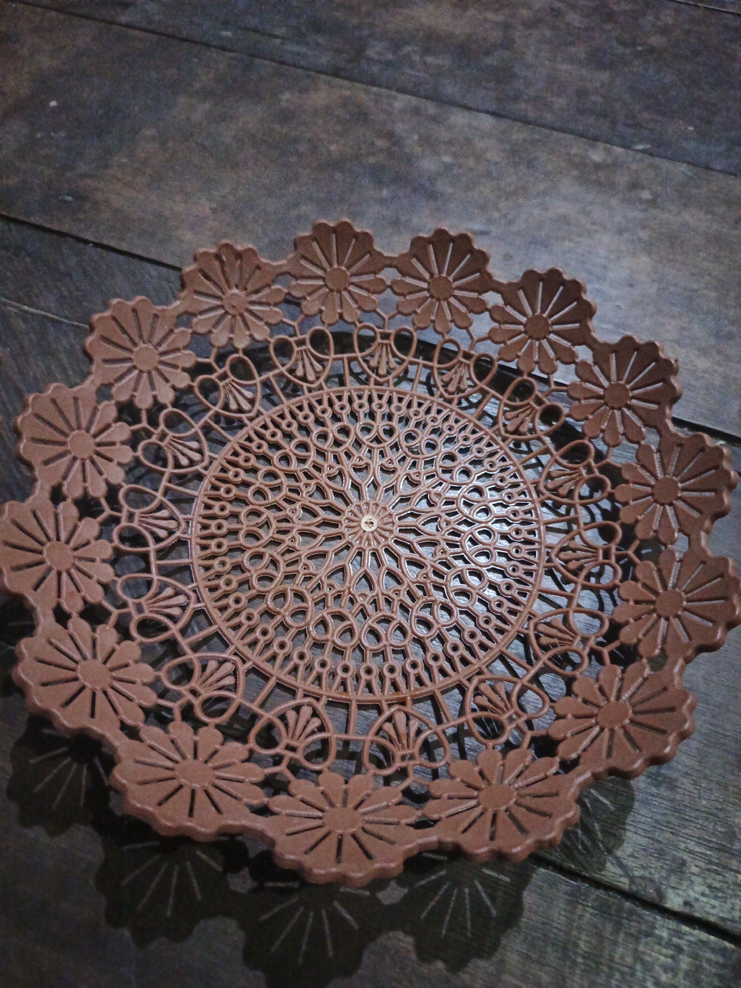 The image shows a brown, plastic, doily-like serving tray with a floral and circular lace-like pattern. The tray is circular with a scalloped edge, each scallop featuring a flower design. The interior of the tray has intricate, symmetrical patterns. The tray is placed on a dark wooden surface, which appears to be a table or floor with visible wood grain. Shadows from the tray are cast on the wooden surface, indicating a light source from above and to the right. The background is dark and out of focus, with hints of wood grain. The image appears to be an indoor still life photograph. There are no people or animals visible in the image. The location is described as the city of Sinar Bulan, Indonesia.