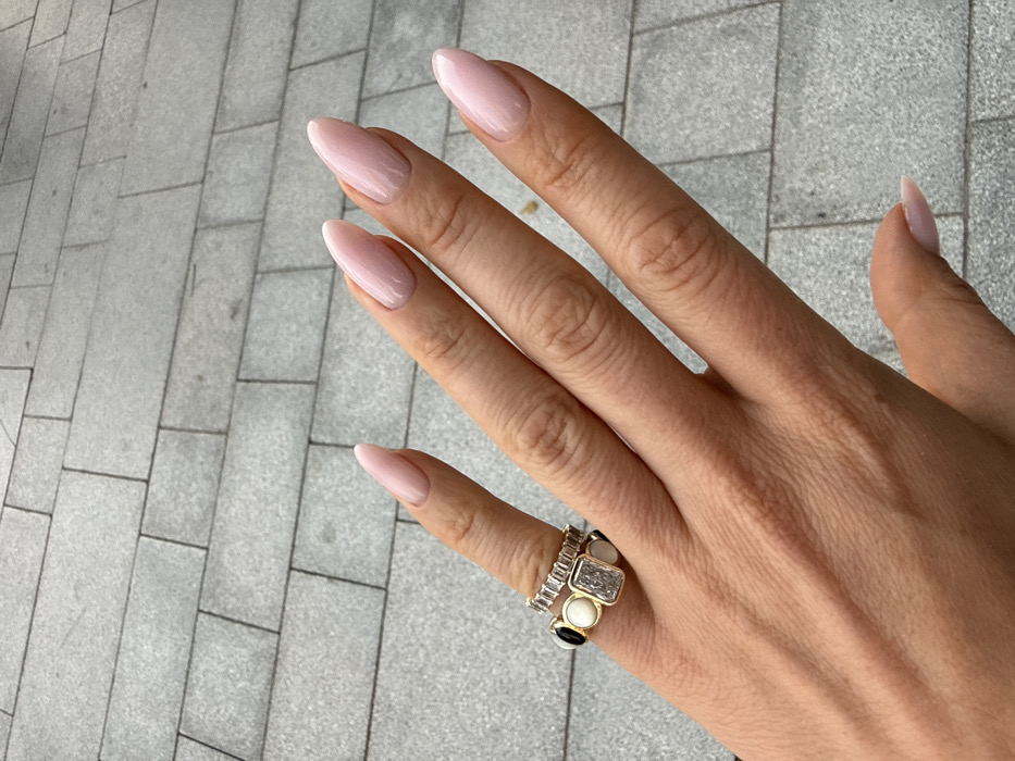A close-up image displays a light-skinned left hand positioned over a paved surface. The hand exhibits five fingers, all featuring elongated, almond-shaped fingernails coated with a glossy, light pink or nude polish. Two rings adorn the ring finger: an upper gold-toned eternity band set with numerous baguette-cut clear stones, and a lower wider gold-toned band featuring alternating black and off-white circular inlays, centered by a prominent rectangular clear stone. The background consists of a grey pedestrian paving made of rectangular blocks arranged in a staggered pattern. No specific landmarks or environmental features are present to definitively identify the location within Dubai, United Arab Emirates.