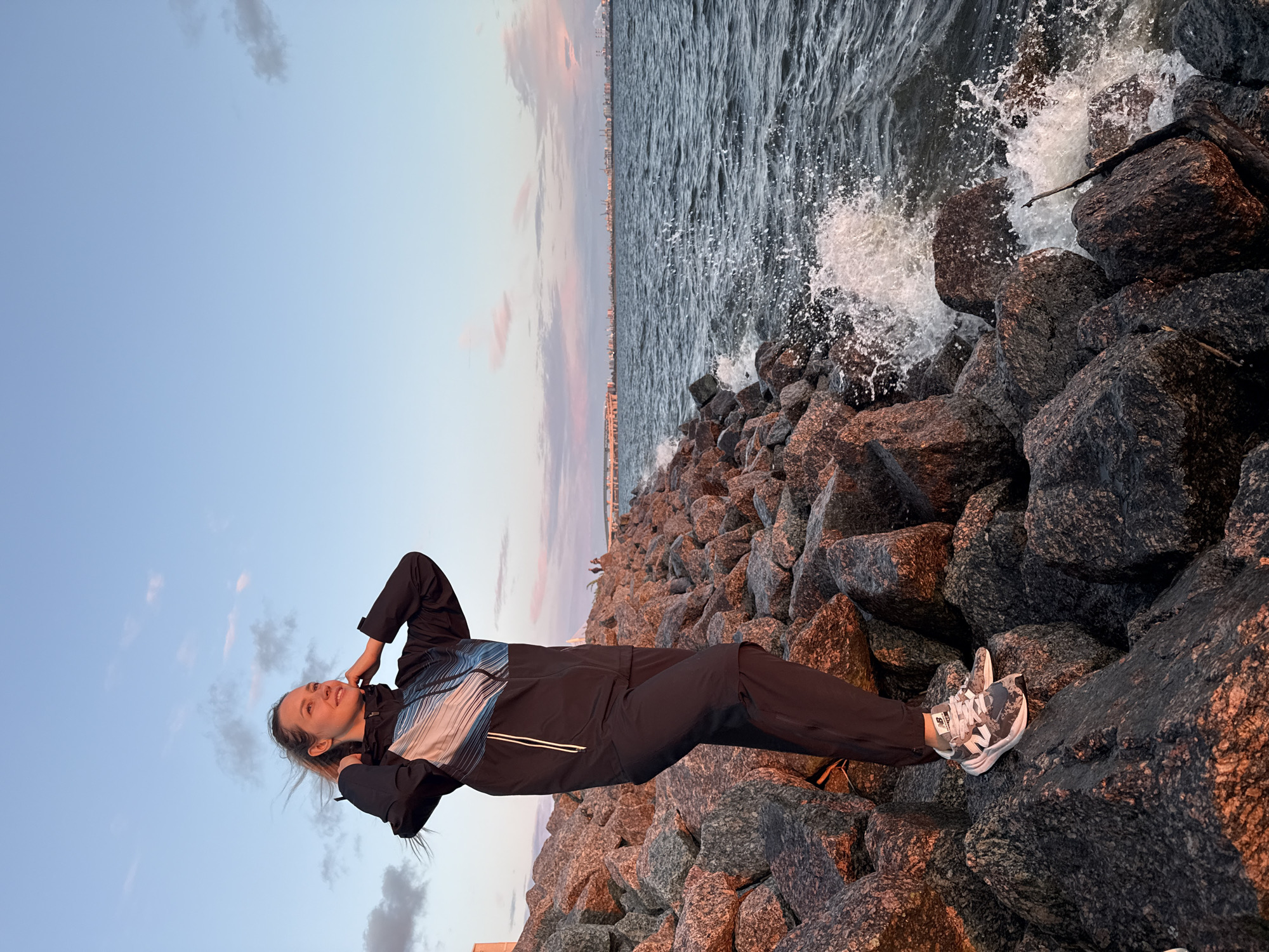 A woman stands on a rocky shore, adjusting her jacket. The sky is a pale blue with streaks of pink and orange clouds, suggesting either sunrise or sunset. The sea is dark and choppy, with waves crashing against the rocks. In the distance, a city skyline is visible, with buildings and cranes indicating an industrial or port area. The woman is wearing athletic clothing, including a dark jacket with a blue and white pattern, dark pants, and athletic shoes. Her hair is blonde and windswept. The rocks are large and uneven, appearing to form a breakwater. The overall impression is one of a peaceful yet dynamic natural setting within an urban environment. The location is likely the Gulf of Finland near Saint Petersburg, Russia, given the characteristic urban landscape and seascape.