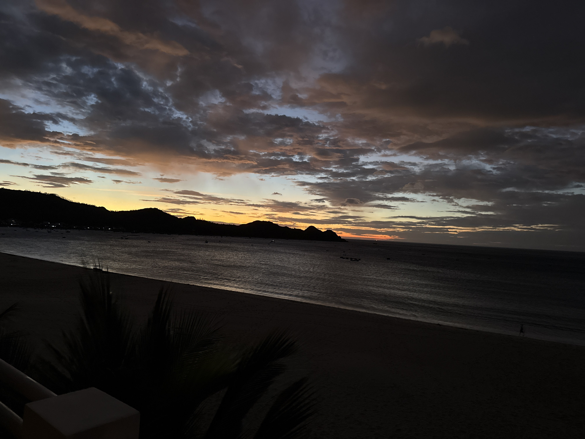 The image shows a sunset over a beach in Song Cau, Vietnam. The sky is filled with dramatic, dark clouds with streaks of orange and yellow from the setting sun. Silhouetted hills line the horizon, and the ocean is dark and calm, with small waves lapping at the shore. A dark, sandy beach stretches into the foreground, with palm fronds casting shadows. In the distance, a few small boats are visible on the water, and lights can be seen along the coastline. The overall mood is serene and tranquil.