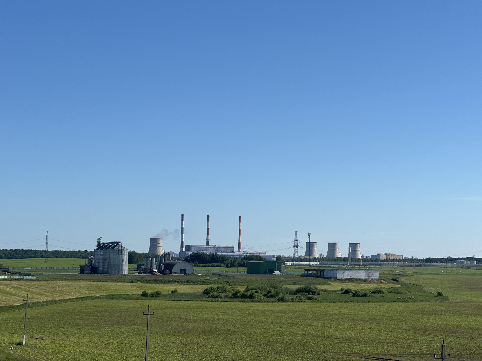 This media file captures a wide-angle view of an industrial complex situated in a rural landscape. The scene is dominated by a clear, bright blue sky, suggesting fair weather and daytime. In the mid-ground, a power plant is visible, characterized by several tall, red and white striped chimneys emitting plumes of white smoke, and large, concrete cooling towers with vapor rising from them.
To the left of the power plant, a large, cylindrical metal grain silo stands prominently, accompanied by smaller industrial structures. The industrial site is surrounded by green fields, some appearing cultivated and others more wild with scattered bushes. In the foreground, the green fields stretch out, with power lines and simple wooden utility poles dotting the landscape. A few low-lying, elongated green buildings are also part of the industrial complex, along with some white rectangular structures that might be storage or processing units.
The overall impression is one of an active industrial zone within a predominantly agricultural or undeveloped area. The sunlight suggests it is likely mid-morning or early afternoon. There are no people or animals visible in the frame, focusing the attention solely on the industrial structures and the natural surroundings. The location is identified as Шчомысліцкі сельскі Савет, Belarus, which provides geographical context for this scene of energy production within a Belarusian rural setting. There is no visible text in the image.