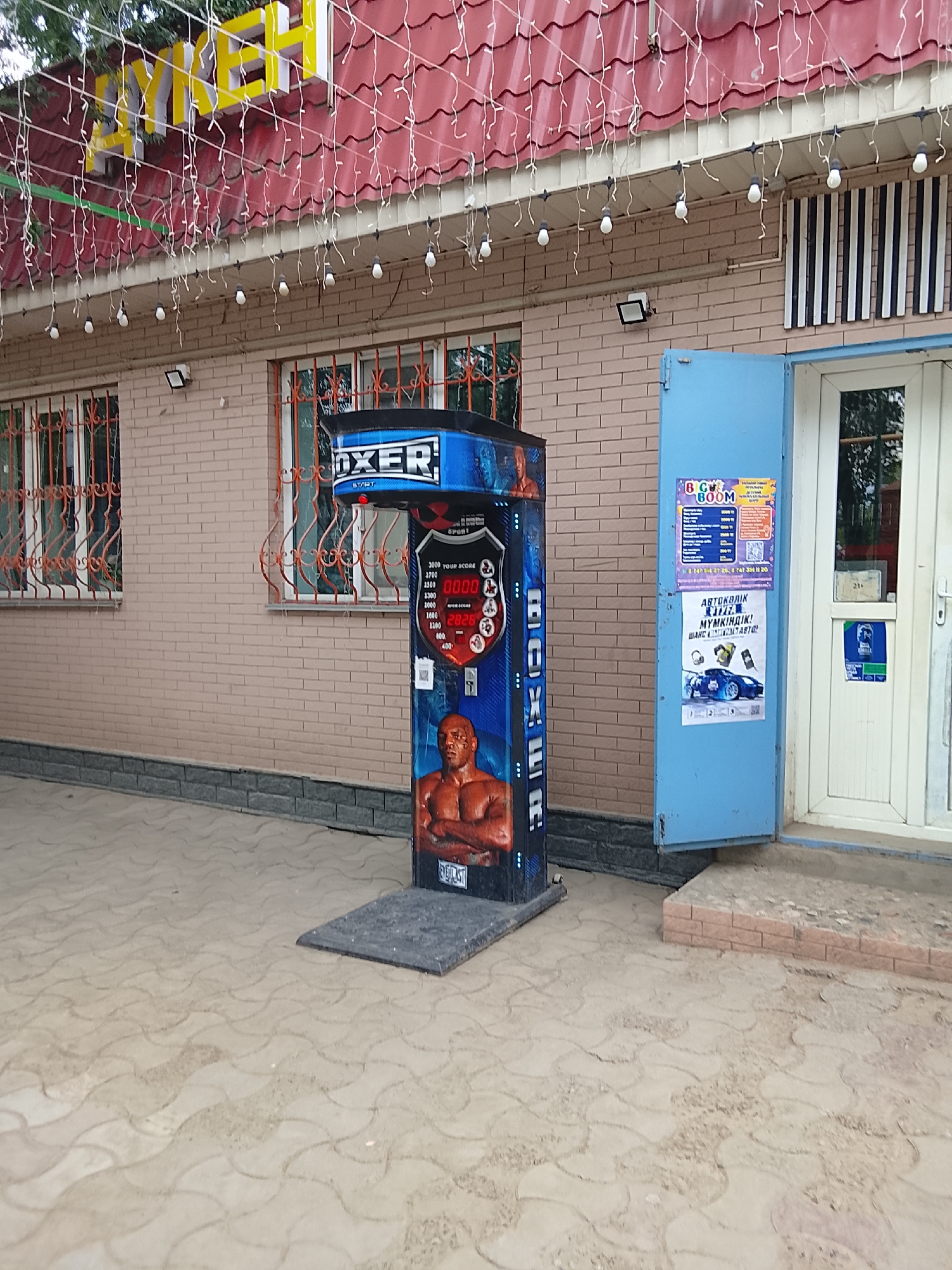 The image shows a street scene in the city of Жалпақсай, Kazakhstan. In the foreground, a punching bag game machine with a depiction of a boxer is positioned on a patterned paved surface. The machine features a digital display showing "00.00" and "HIGH SCORES" with the numbers "2826" visible. To the right of the punching bag machine is a bright blue door with posters on it. One poster advertises "CAR BOOM" with prices and contact information, and another features an image of a blue sports car with text related to car parts and sales. The building behind the machine has brick walls and windows with decorative orange metal bars. Above the building, a red tiled roof is visible, adorned with strings of white lights. The overall environment appears to be an outdoor commercial or entertainment area.