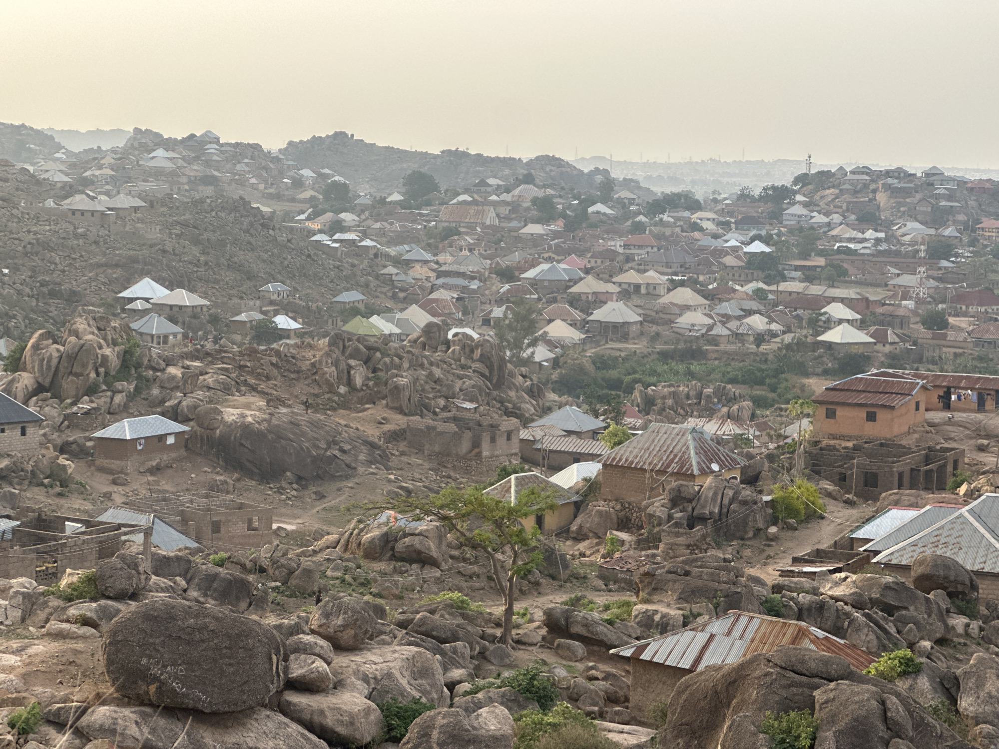 The image shows a dense settlement built among large rock formations, likely located near Jos, Nigeria. The area is characterized by clusters of buildings with corrugated metal roofs, interspersed with large boulders and sparse vegetation. The structures appear to be a mix of completed homes and partially constructed buildings. The terrain is rugged and rocky, with the buildings seemingly integrated into the natural landscape. The overall scene is somewhat hazy, with the background buildings appearing more distant and less defined. The architecture is simple and functional, reflecting the local building style.
