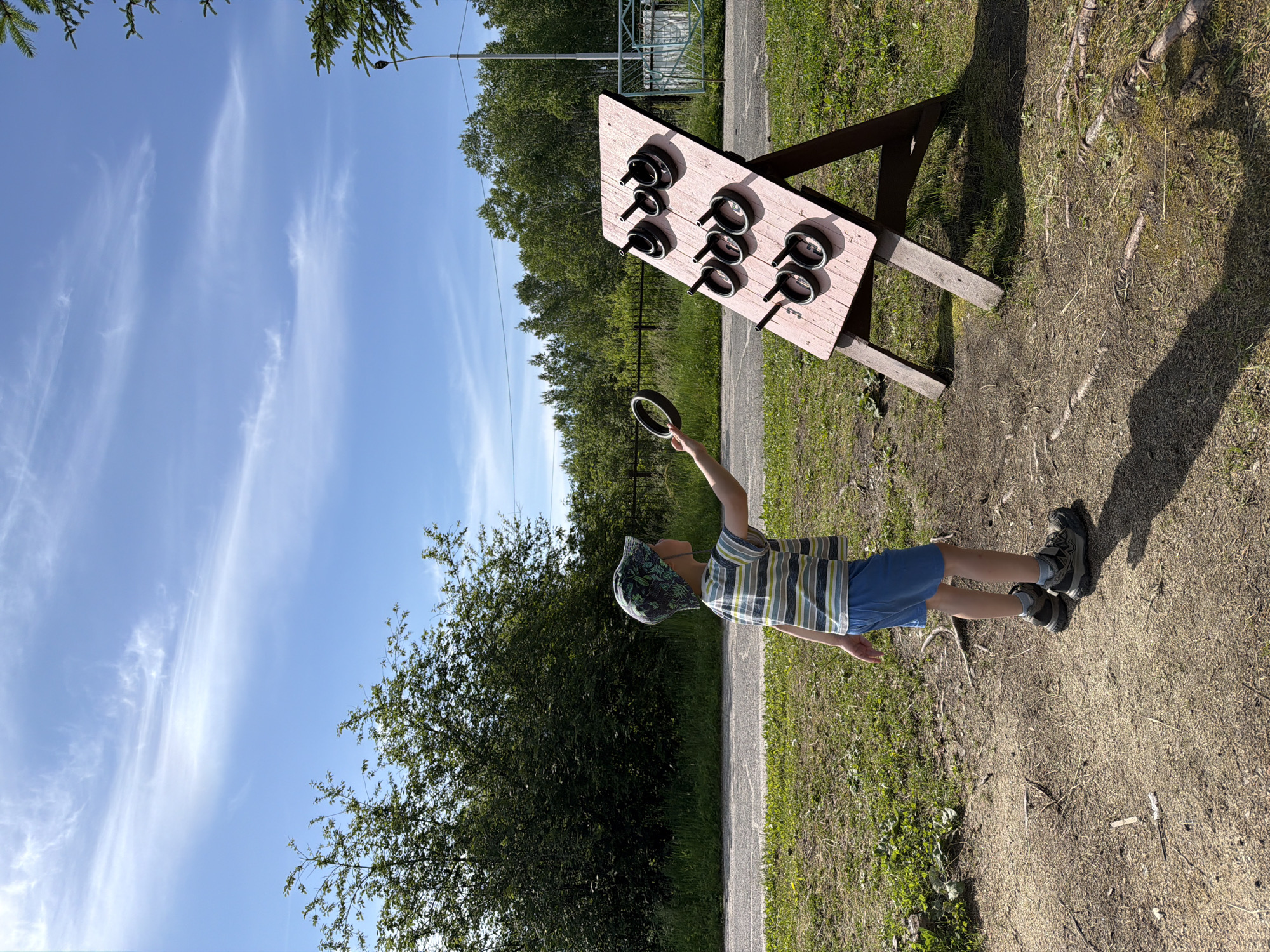 A young boy, facing away from the camera and towards a ring toss game, stands in a grassy, outdoor area under a bright blue sky with wispy clouds. He is wearing a striped t-shirt, blue shorts, a patterned cap, and hiking shoes. He holds a dark ring in his right hand, poised as if to throw it at the game. The ring toss game is a wooden board, painted pink, propped up on two A-frame wooden legs. It has nine black pegs arranged in three rows, designed to catch the thrown rings. The background features a dense line of green trees, a paved path, and a metal fence. The ground is a mixture of dirt and grass, with some scattered twigs. A lamppost is visible on the right side of the image. The scene is likely located in an outdoor recreational area or park in Kul'dur, Russia.