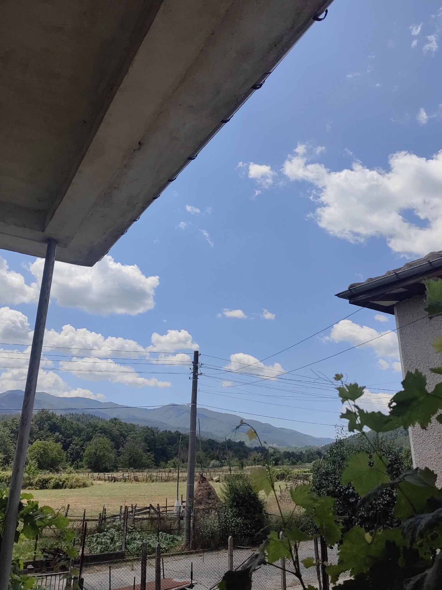 The image depicts a rural landscape under a bright blue sky with scattered white clouds. In the foreground, to the left, a concrete overhang supported by a metal pole juts into the frame, creating a shadowed area. To the right, the corner of a building with a tiled roof is visible. Beyond these structures, a field stretches out, bordered by a rustic wooden fence. Further in the distance, a dense forest covers rolling hills, which in turn give way to more distant, hazy mountains. A few utility poles with power lines are interspersed throughout the scene, indicating a rural settlement. A haystack stands near one of the poles. In the immediate foreground, some leafy green vines are visible, partially obscuring the view of the field and fence. The scene appears to be located in a village like Djerovo, Bulgaria, given the architectural style and the natural environment. No people or animals are present in the image.