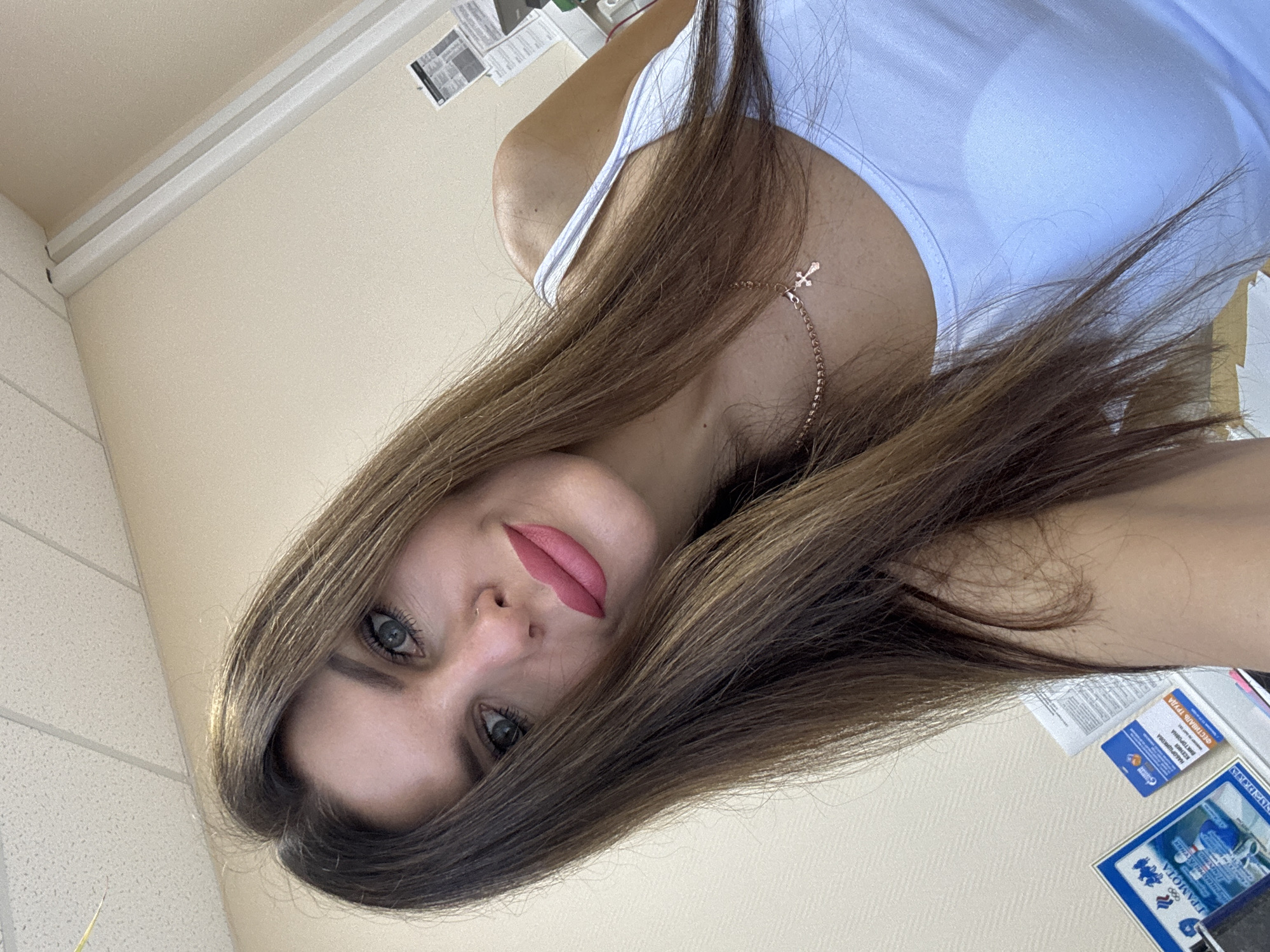 A woman with long brown hair and blue eyes is captured in a selfie. She is wearing a white tank top and a rose gold necklace with a cross pendant. Her lips are painted with pink lipstick. The background appears to be an office environment, with a textured wall and ceiling tiles. There are some papers and documents visible on a surface in the lower left and right of the image. The image was taken in Chaikovsky, Russia.