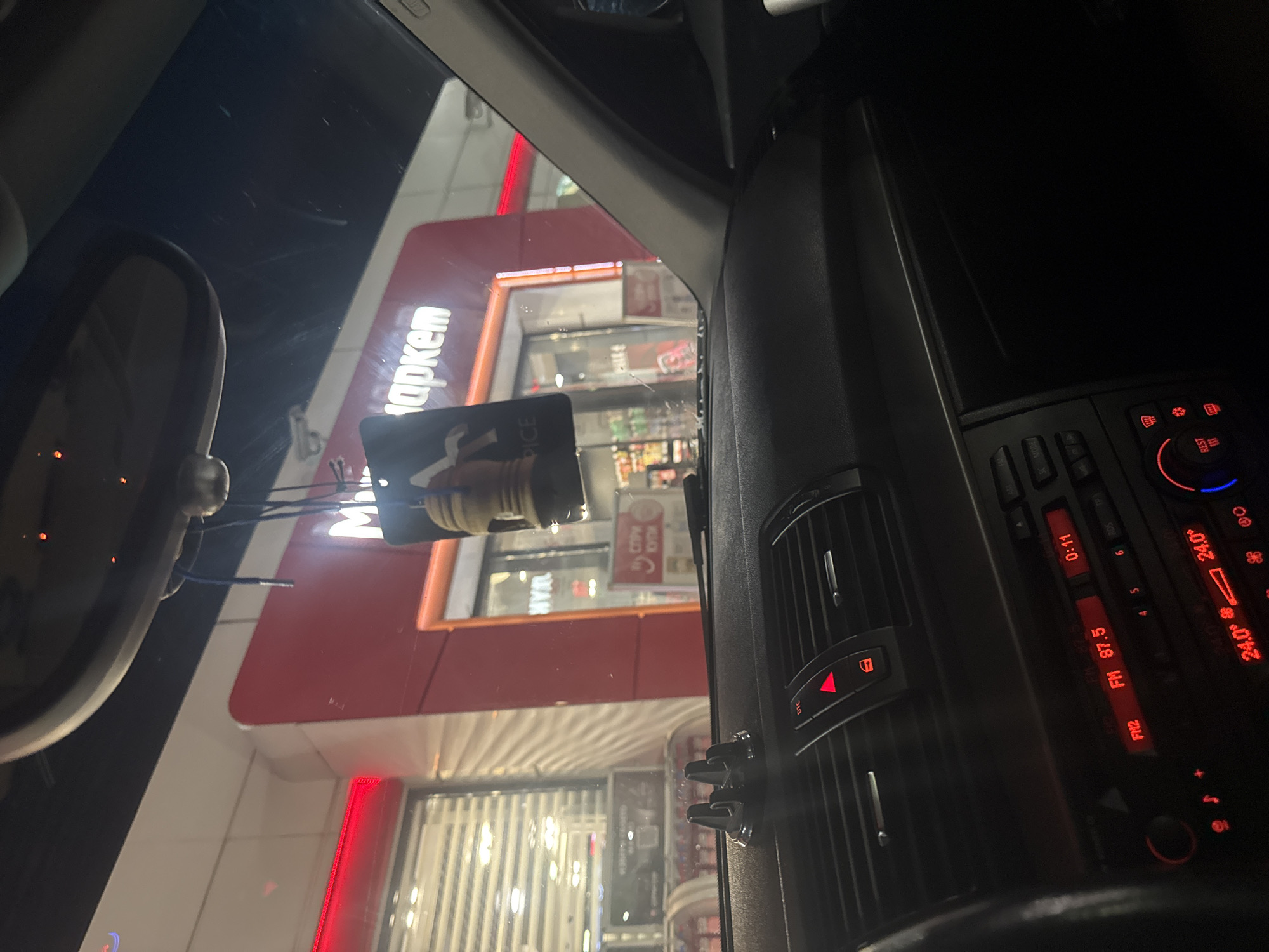 The image is taken from inside a car, looking out through the windshield at what appears to be a "Маркет" (Market) store. The store has a red and white exterior with glass windows. In the foreground, the interior of the car is visible, including the rearview mirror, dashboard air vents, and the car's radio/climate control panel. The radio display shows "FM 87.5" and a temperature of "24.0°". A card is hanging from the rearview mirror. The environment appears to be an urban or commercial area. The image does not include specific details to confirm the location as Vodenichane, Bulgaria. There are no people visible in the image.