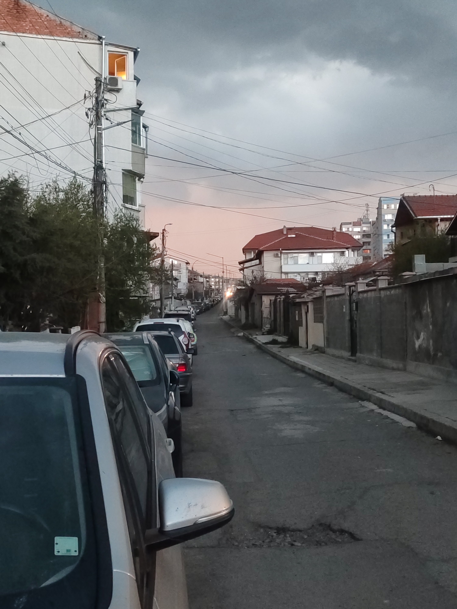 The image depicts a quiet street in Burgas, Bulgaria, at dusk or early evening. The sky is largely overcast with dark grey clouds, but a subtle pinkish-orange hue brightens the horizon, indicating twilight.
On the left, a row of parked cars extends down the street, with the front section of a silver vehicle dominating the foreground. A small, green and white sticker, likely a vehicle permit, is visible on its windshield, though any text is unreadable. Behind the cars, a tall, white building with a red-tiled roof rises, featuring a brightly illuminated top-floor window that emits a warm, yellow light. A dense network of black utility wires is strung across the frame, anchored by a wooden pole next to the building. Lush green trees and bushes grow along this side.
The right side of the street features a long, unadorned grey concrete wall, bordering residential properties with red-tiled roofs. Beyond these, more buildings with similar architectural styles line the street. In the distant background, taller, modern apartment blocks are discernible against the fading light, some with visible illuminated windows.
The asphalt road shows considerable wear, with a prominent pothole in the foreground. A concrete sidewalk and curb run along the right edge. The scene is devoid of people or discernible activity, lending a calm and somewhat introspective mood. The blend of traditional tiled roofs with modern high-rises and abundant overhead wiring defines this urban residential setting.
