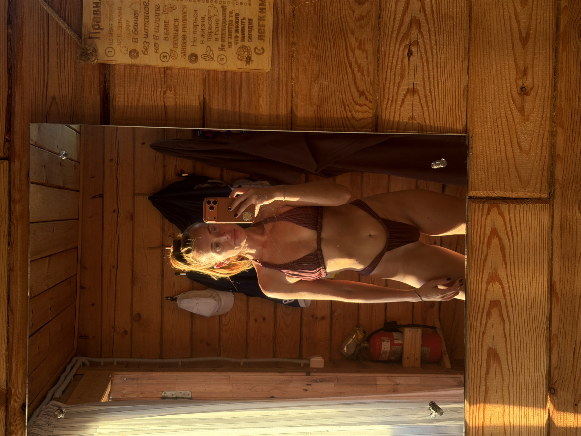 This media file shows a mirror reflection of a woman in a bikini, standing in a room with wooden walls. The room appears to be a sauna or a similar rustic structure, with exposed wooden planks for walls and a wooden ceiling. Sunlight streams in from the left, creating a warm, bright atmosphere and casting shadows.
The woman is looking at her phone, which she is holding to take the selfie. She has blonde hair and is wearing a dark-colored bikini. Her body is facing the mirror, and her gaze is directed at her phone's screen.
In the reflection, we can see the room's interior behind the woman. There are various objects on the wooden shelves and walls, including a fire extinguisher, what appears to be a white baseball cap, and some plumbing pipes. A string hangs from the ceiling on the left side, attached to a wooden sign with Russian text.
The visible text on the wooden sign, read from right to left, appears to be a list of rules or advice related to saunas. The words "Прави" (Rules) are visible at the top, followed by numbered points with instructions and phrases such as "В баню" (To the bathhouse), "помылся - заново родился" (washed - reborn), and "Не откладывай на завтра то, что можно вымыть но сегодня" (Don't postpone until tomorrow what can be washed today).
The overall mood is relaxed and casual, with the natural lighting and rustic setting suggesting a comfortable and perhaps private moment. The time of day is likely daytime, given the strong sunlight.