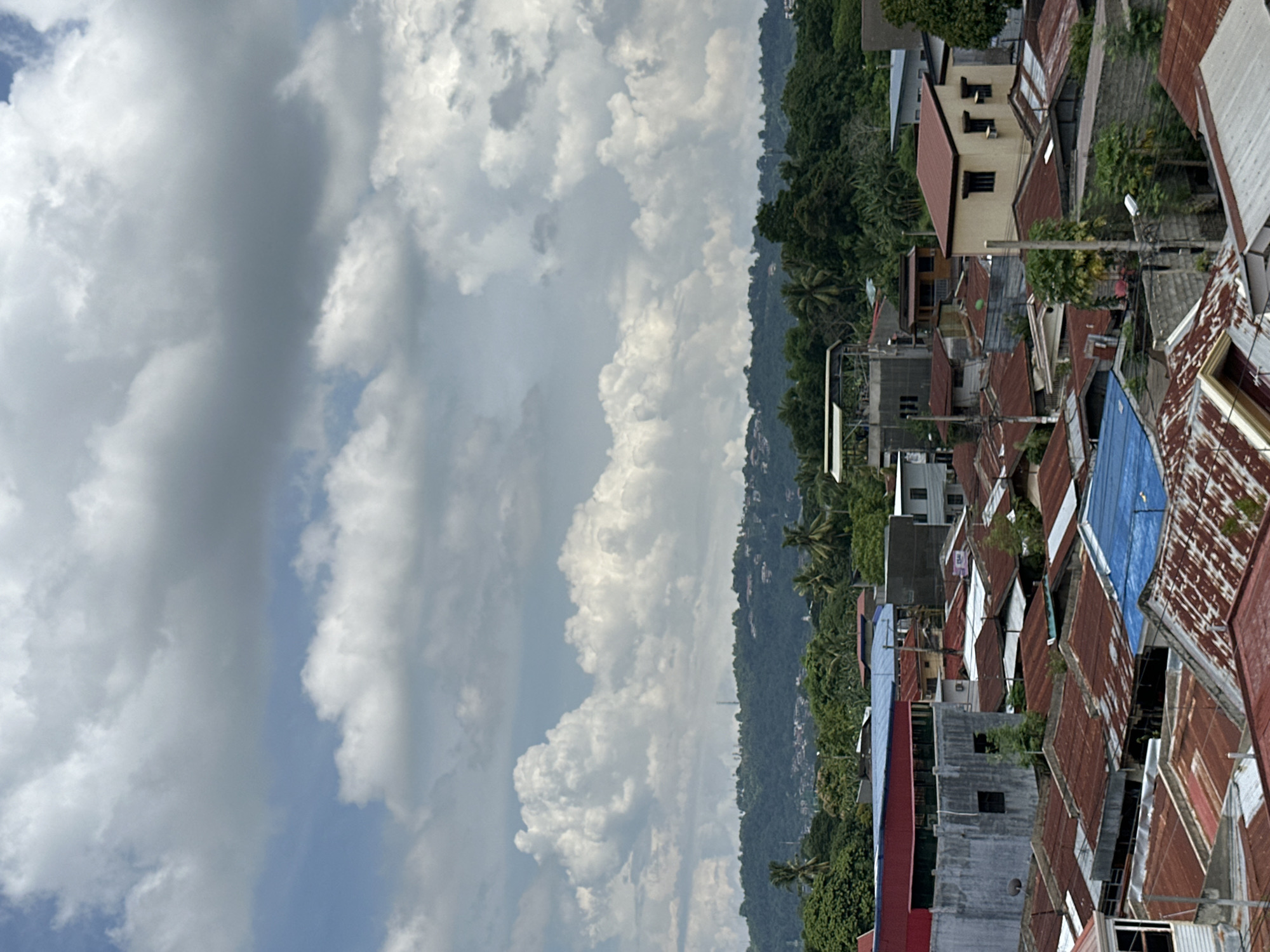 The image captures a scene from Davao City, Philippines. The foreground features densely packed buildings with corrugated metal roofs, predominantly red and blue. A forest stretches in the middle ground, and in the distance, heavy white clouds fill most of the sky.