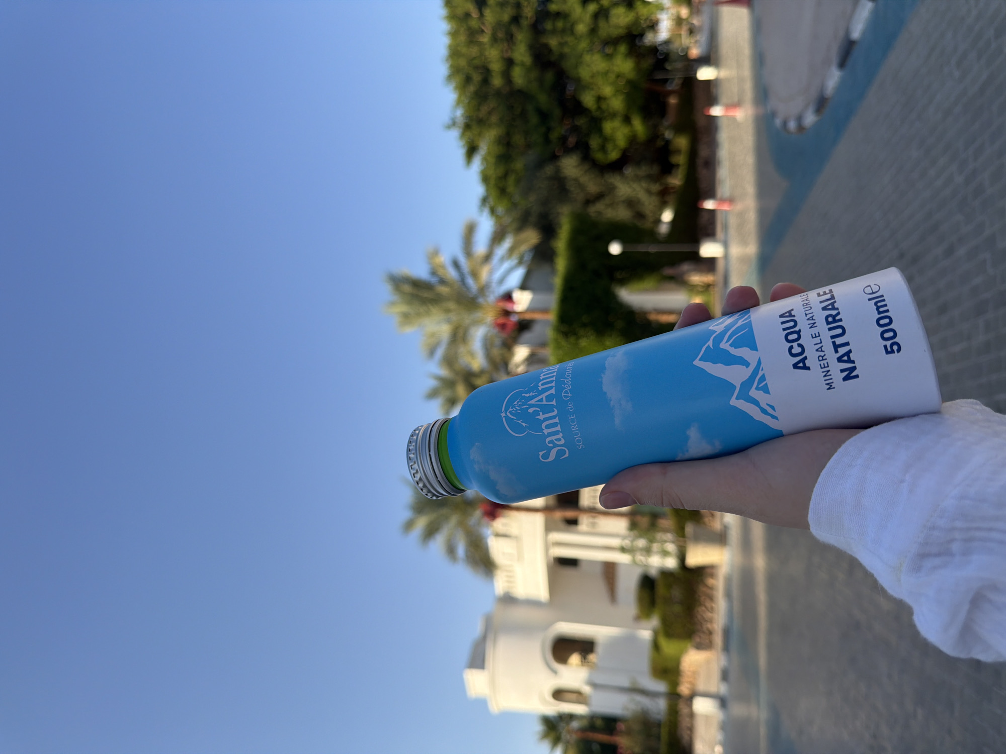 A hand wearing a white shirt is holding a light blue metal water bottle against a clear blue sky. The bottle has white text and graphics, including a mountain range and clouds. The text on the bottle reads "Sant'Anna SOURCE de Pedoures ACQUA MINERALE NATURALE 500mlE". In the background, a white building with arched windows and lush green trees are visible, suggesting a resort or hotel in a warm climate, possibly Sharm El Sheikh, Egypt, given the palm trees and arid architectural style. The ground in the foreground appears to be a paved pathway with blue lines and a curb.