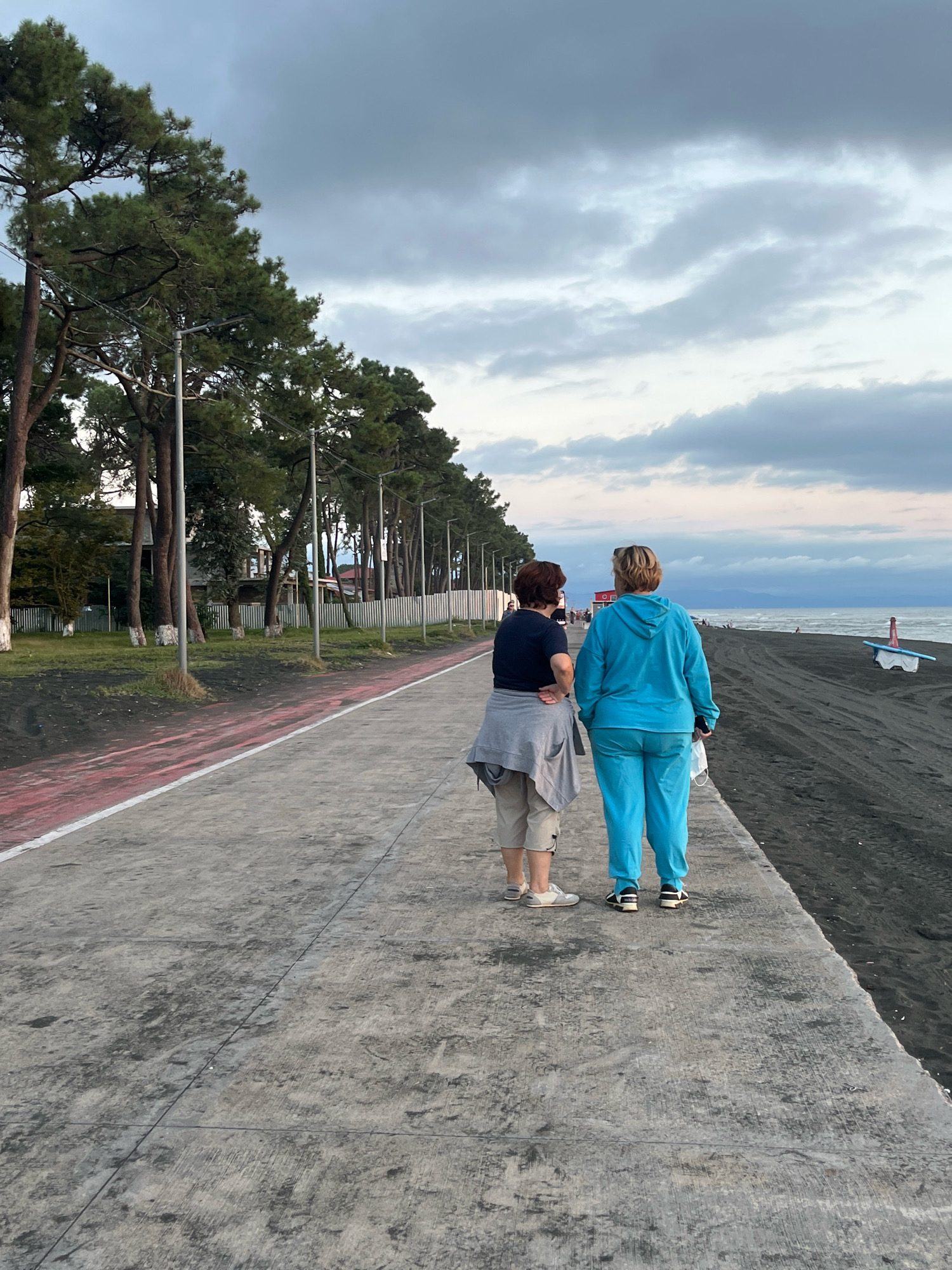 Two individuals, appearing to be women, stand facing away from the camera on a concrete promenade. The woman on the left wears a gray cardigan over a dark t-shirt and tan shorts, with white sneakers. The woman on the right is dressed in a blue tracksuit and white sneakers, holding a white mask in her right hand. They are positioned on a wide, gray concrete promenade which has a red stripe along its left edge.
To their left, a grassy area is visible, lined with tall evergreen trees. Several lampposts are spaced along this tree line. Beyond the trees, a white metal fence separates the area from some low-rise buildings.
To their right, a dark sand beach stretches towards a body of water, likely the Black Sea, given the Ureki, Georgia location. The water appears calm, with a faint shoreline visible in the distance. A small, light-colored boat is beached on the sand. The sky is overcast with dramatic clouds, suggesting either early morning or late afternoon light. The overall scene depicts a tranquil coastal promenade setting in Ureki, Georgia.