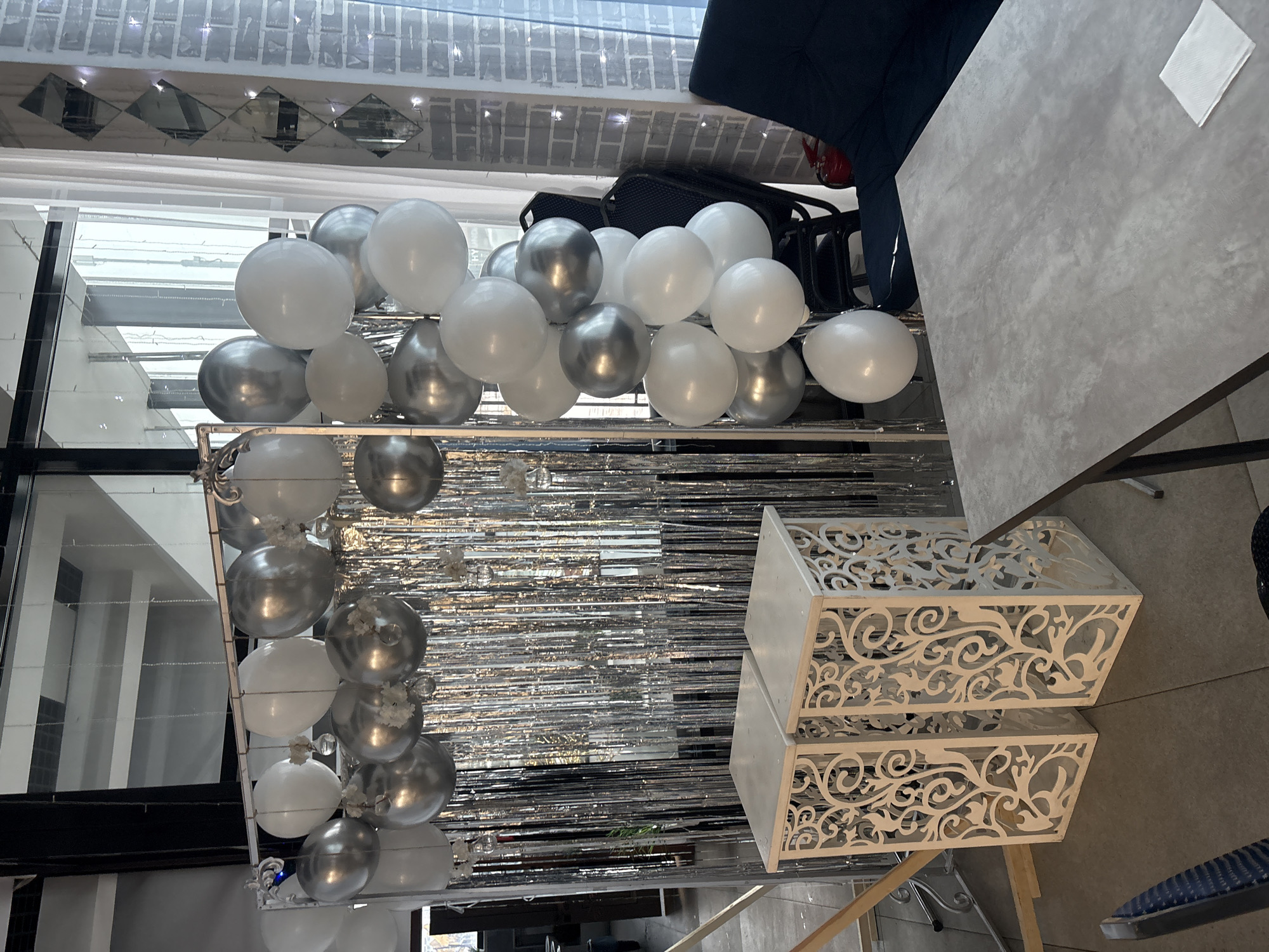 An indoor scene in Solontsy, Russia, showcases an elegant celebration setup, predominantly featuring white and silver decorations. Numerous round balloons, alternating between opaque white and reflective silver, are arranged, possibly forming an arch or garland, with some smaller clusters attached to a decorative structure. Behind these balloons, a shimmering backdrop of silver metallic tinsel or foil streamers hangs, adding to the festive atmosphere.
To the right, two white, ornate decorative boxes are stacked. These boxes feature intricate, laser-cut patterns with swirling, organic designs, contributing a touch of sophistication. A grey, textured tabletop is visible in the upper right, with a neatly folded white napkin placed upon it. Part of a dark blue upholstered chair or seat, along with black chair frames, can be seen near the table.
The background reveals architectural details, including what appears to be a wall or ceiling adorned with square and diamond-shaped mirrored tiles. Large windows or glass panels with reflections are also visible, suggesting a brightly lit space. The overall color scheme is monochromatic, with shades of white, silver, and grey, creating a chic and celebratory ambiance. The scene is well-lit, likely a combination of natural and artificial light. No visible text is present in the image.