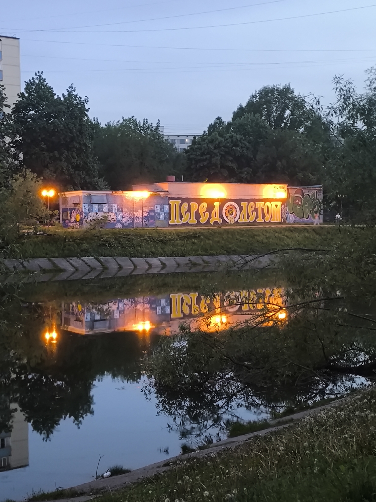 The image shows a building with graffiti in an urban setting in an unknown city. The graffiti is brightly lit and reflected in the water in front of the building. The surrounding area includes trees, grass, and a paved path. In the background, other buildings are visible, and a person is seen riding a bicycle near the graffiti-covered building. The lighting indicates it is either dusk or dawn.
