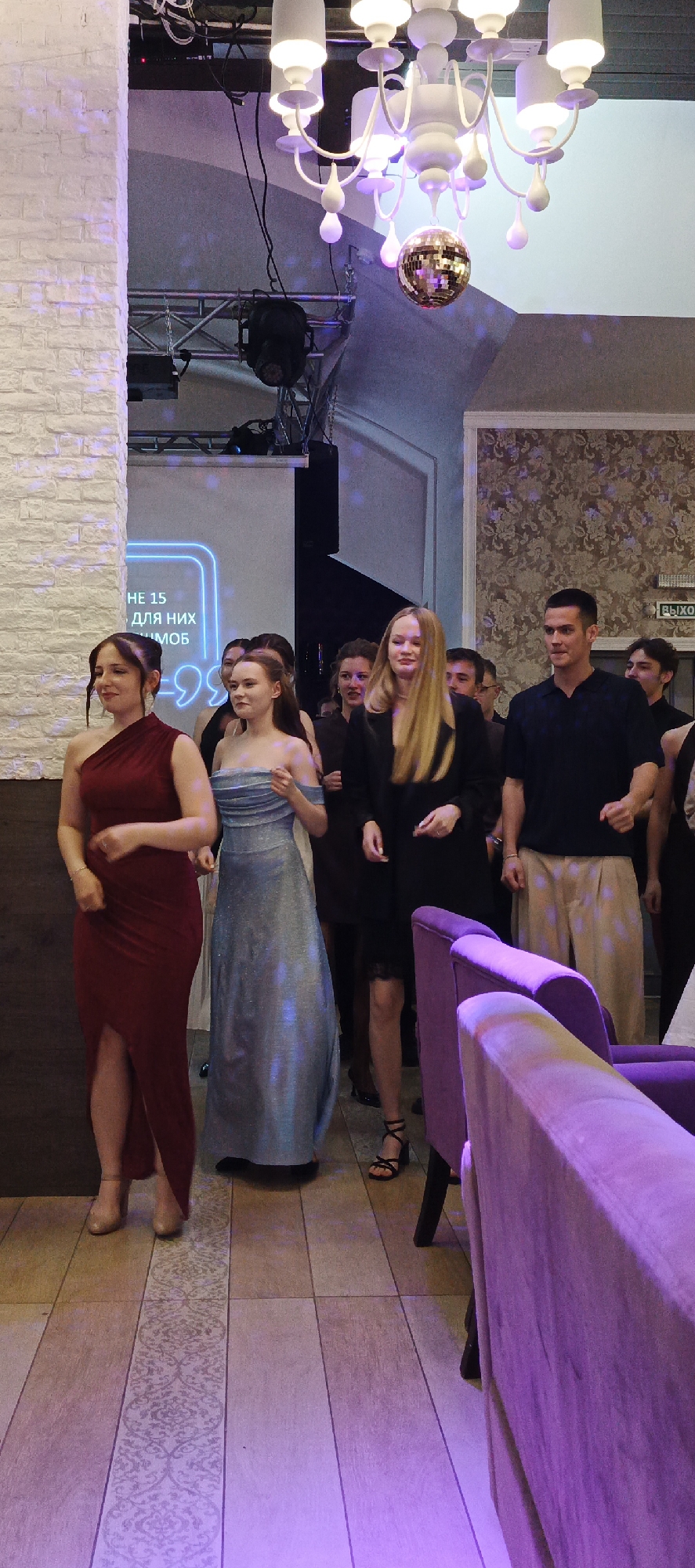 The image captures an indoor event, possibly a formal gathering or celebration, with a group of young adults present. In the foreground, three young women are prominently featured. The woman on the left wears a floor-length, deep red, one-shoulder gown with a high slit. Next to her, a woman in a shimmering, light blue, off-the-shoulder gown is visible. To the right of the women, a young man in a dark blue collared shirt and light-colored pants is standing, appearing engaged in conversation or activity. Behind them, a larger group of individuals can be seen, some facing the camera, others looking away.
The environment suggests a decorated space, with a white brick wall on the left, and a patterned wallpaper on the right. A large chandelier with multiple lights and a disco ball hangs from the ceiling, hinting at a festive atmosphere. Lighting appears to be a mix of ambient and colored, with purple hues prominent in certain areas, particularly on the floor and furniture. In the background, a screen displaying text is partially visible, with the words "НЕ 15" (not 15) and "ДЛЯ НИХ" (for them) discernible. The flooring is tiled with a decorative border. To the far right, a sign with the Russian word "ВЫХОД" (EXIT) is mounted on the wall, indicating an exit.
The image's metadata may indicate a location in Chaikovsky, Russia, given the presence of Russian text within the scene. The overall impression is one of a lively event, with young people dressed for an occasion.