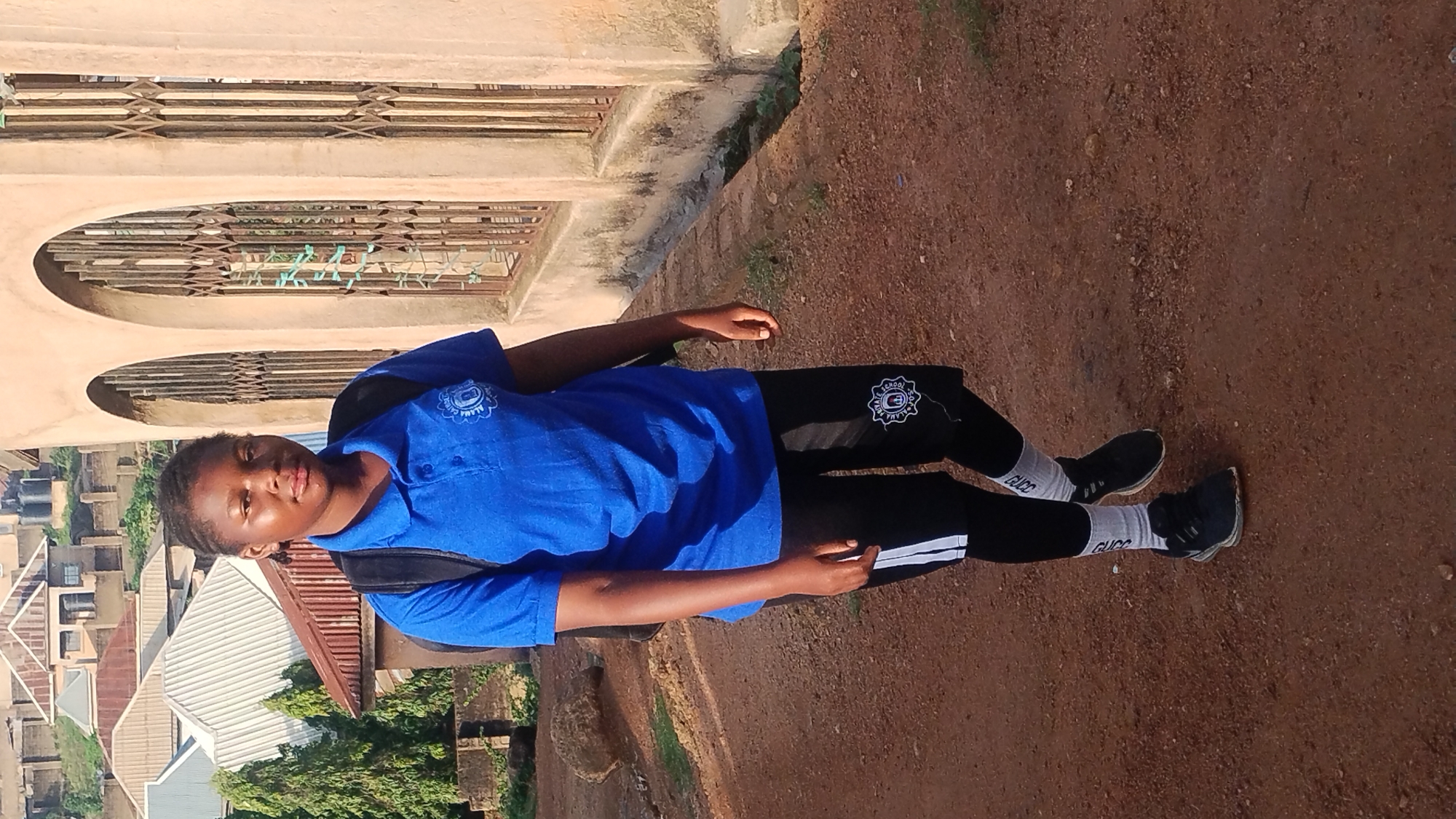 The image displays a young individual standing outdoors on a reddish-brown dirt ground. The person, who appears to be female with dark skin and short dark hair, is looking directly at the camera with a calm, neutral expression. She is wearing a bright blue short-sleeved polo shirt with a circular emblem on the left chest, which contains text that is partially legible, possibly "ALAMA SCHOOL" or similar. She also wears black athletic pants featuring a similar circular emblem on the upper left thigh. Her feet are clad in white ankle socks with the text "GUCC" (likely "GUCCI") visible, and dark sneakers. A black backpack is on her back.
In the background, there is a light-colored building with several arched openings or windows fitted with horizontal metal grates. Further beyond, residential buildings with various rooftops and some green trees are visible, suggesting an urban or semi-urban setting. The lighting is bright and clear, indicating it is daytime under sunny skies.