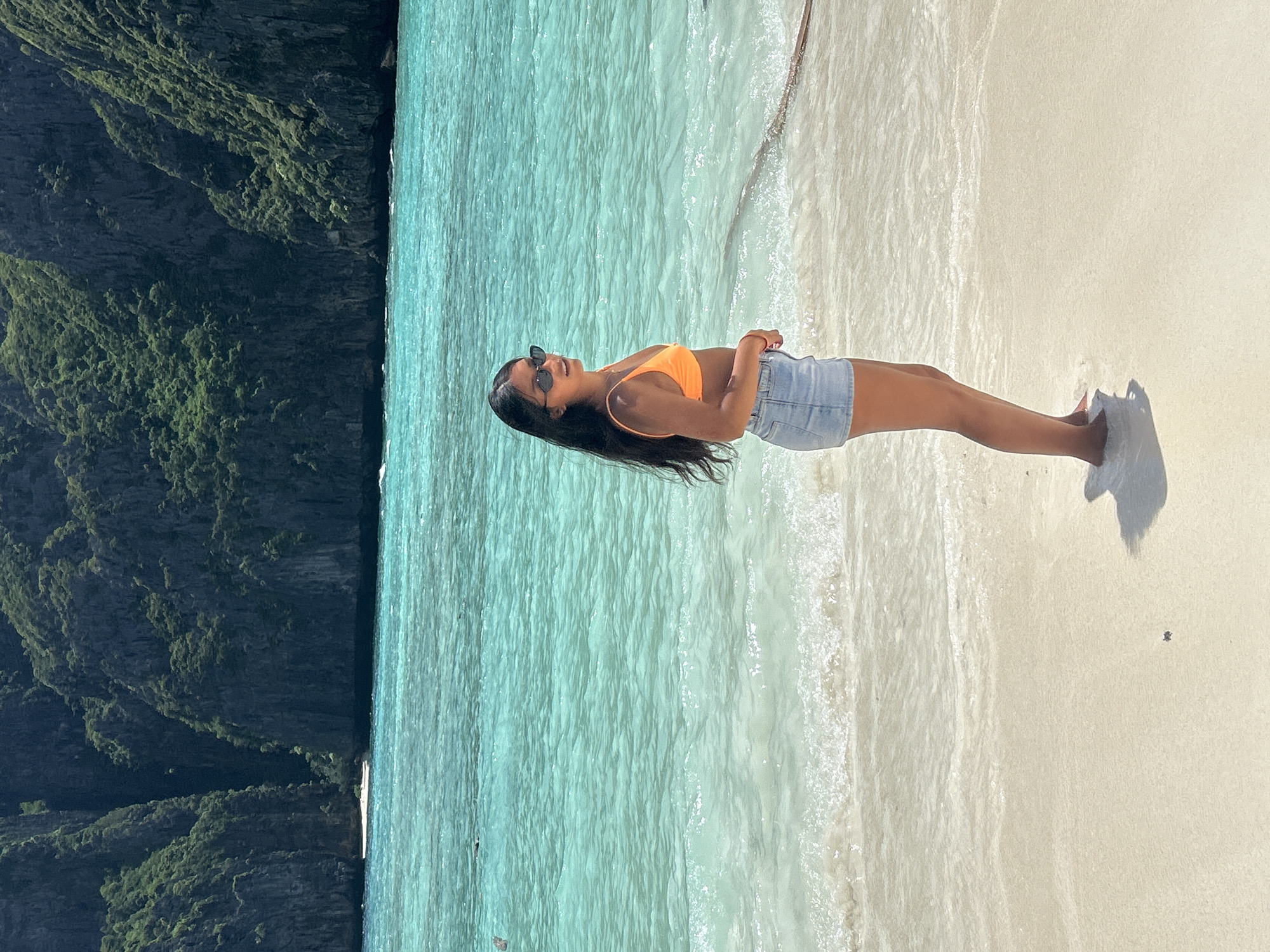 A young woman stands on a white sand beach with clear turquoise water lapping at her feet. She is wearing an orange bikini top and light blue denim shorts. Her long, dark hair is down and she is wearing sunglasses. The water is calm and appears shallow near the shore, with gentle waves. In the background, large, green, rocky cliffs rise dramatically from the sea. A thin rope is visible in the water behind her, stretching towards the right side of the frame. The scene is brightly lit by sunlight, casting a shadow of the woman on the sand. This appears to be Maya Bay, Phi Phi Leh island, a popular tourist destination located within the Ao Nang district of Krabi province, Thailand.