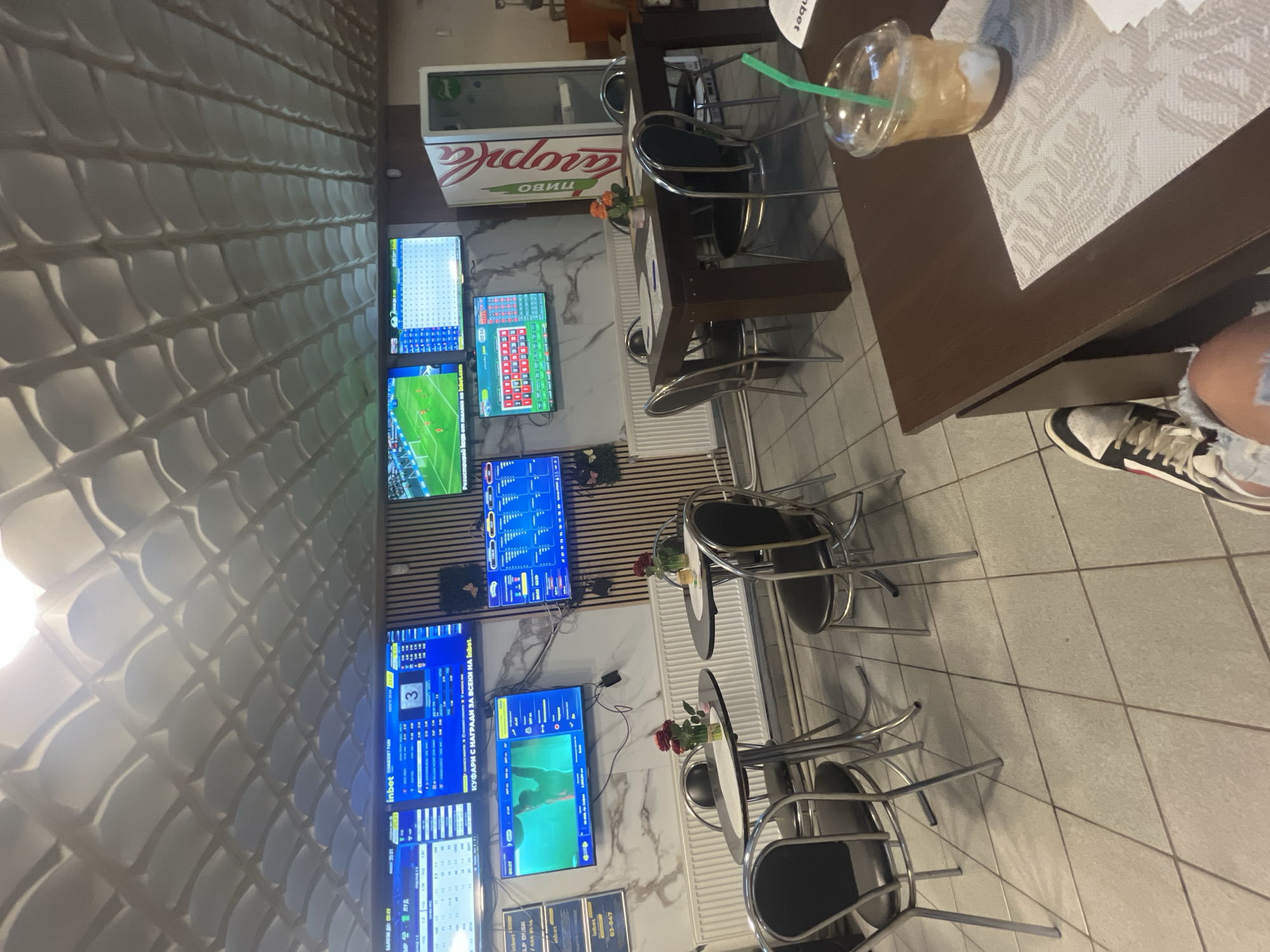 The image shows the interior of a sports bar or betting shop, likely located in Batoshevo, Bulgaria. The walls feature textured decorative panels, and numerous television screens display live sports events, possibly soccer, and betting information. Several small tables with metal chairs are arranged in the room, suggesting a casual seating layout. A plastic cup with a straw and some paperwork is visible on one of the tables, alongside part of a person's leg with sneakers. There is a refrigerator behind one of the tables.