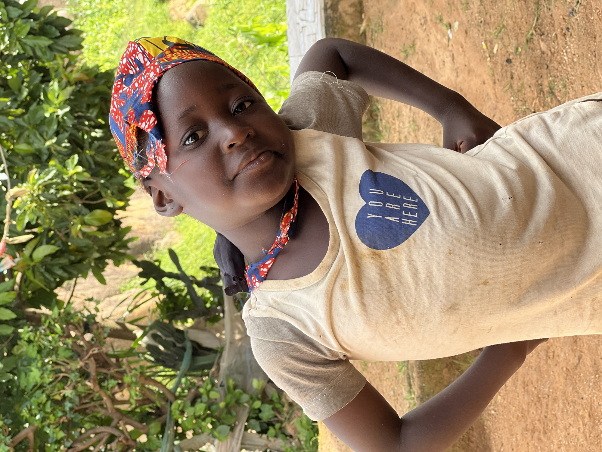 This is a close-up, portrait-style photograph of a young Black child wearing a colorful headscarf and a light-colored t-shirt. The child is looking directly at the camera with a calm expression. The t-shirt has a blue heart graphic on it with the text "YOU ARE HERE" inside. The background is blurred, showing greenery and what appears to be a dirt path or ground. The lighting suggests it is daytime, possibly late morning or early afternoon, with natural light illuminating the subject. The child's skin tone is dark, and they have subtle red markings on their cheeks, potentially face paint or natural skin marks. The overall impression is that of an informal, outdoor portrait taken in a rural or semi-rural setting, consistent with the location context of Jos, Nigeria.