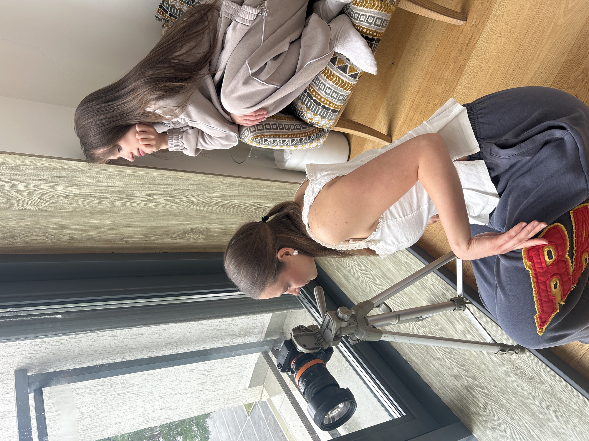The image shows a scene inside a building, potentially a home or studio, in Izhevsk, Russia. A young woman with her hair in a ponytail is setting up a camera on a tripod near a window. The camera is a DSLR with a large lens. Outside the window, a glimpse of greenery suggests a possible park or street with trees. Seated behind her, a younger girl with long brown hair is looking down, seemingly observing the woman. She's dressed in casual clothing, including a light-colored sweatshirt and sweatpants, and is sitting on a chair with patterned cushions. The room appears to have wooden floors and a light-colored wood-like accent wall.