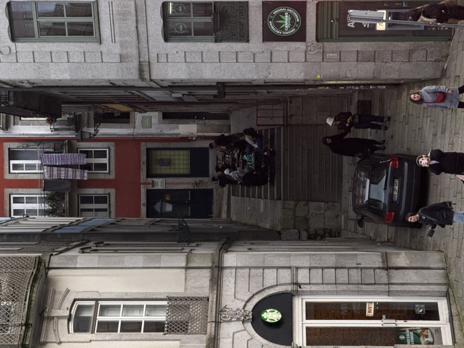 The image captures a vibrant urban street scene, likely an alleyway or a narrow street, lined with multi-story buildings. The perspective is looking down the street, which features a combination of level ground and stone steps. The general architecture suggests an older European city, possibly with Portuguese or Spanish influences, given the tiled roofs and building styles. The lighting appears to be natural daylight, possibly an overcast day or late afternoon, as there are no harsh shadows.
On the left side of the street, in the foreground, stands a light-colored building constructed of what appears to be stone or stucco. The ground floor of this building houses a Starbucks coffee shop, identifiable by its prominent circular green logo featuring a siren against a black, arched facade. The entrance is a large glass door with a large adjacent glass window. Text reading "WELCOME" is visible vertically on the glass door. Further down this side of the street, more traditional building facades are visible.
On the right side of the street, in the foreground, there is another light-colored building with large windows, one featuring a decorative metal grate. Moving further down, a red-painted building stands out, featuring multiple white-framed windows. From one of these windows, several pieces of laundry, possibly striped towels or blankets, are hung to dry on clotheslines, adding a domestic touch to the urban setting. Below the laundry, there is an entrance to a cafe or restaurant, indicated by outdoor seating on stone steps and a decorative, possibly stained-glass, panel above the doorway. Some graffiti is visible on the lower part of the wall near this entrance. Above the red building, another light-colored building continues, and on its upper facade, a prominent square black sign with a circular green logo and text reads "ORIGINAL AMSTERDAM CANNABIS".
The street itself is paved with rough, light-colored stones, suggesting cobblestones or flagstones. In the mid-ground, a series of wide stone steps lead down, where an outdoor cafe is situated. Several people are seated at small tables on these steps, engaged in conversation, suggesting a social atmosphere.
A black Audi car is positioned in the middle of the narrow street, facing away from the viewer. Its license plate, "85 JF 46", is clearly visible.
Several individuals are present in the scene:
* People are seated at the outdoor cafe tables.
* On the steps near the car, a person in a white bucket hat and dark clothing is standing.
* Further down the steps, another person in dark attire is standing.
* To the right of the car (from the viewer's perspective), a woman with a red bag is looking towards the camera.
* Behind the car, slightly to the right, a person in a dark jacket is walking.
* Another person with a striped top is partially visible behind the car, also looking towards the camera.
* A person in a dark jacket with a backpack is visible closer to the Starbucks in the lower right, seemingly walking past.
The scene conveys a bustling yet relaxed urban environment, characterized by traditional architecture, small businesses, and public life.