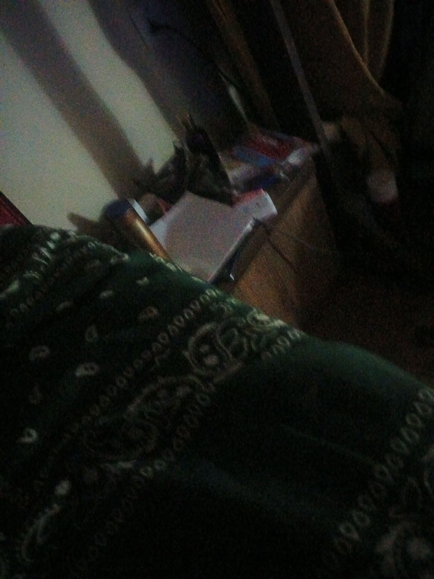 The image depicts a dimly lit indoor scene, likely a room in Jalingo, Nigeria. A dark green bandana with a paisley pattern is prominently featured in the foreground, taking up a significant portion of the frame. To the left and slightly behind the bandana, a wooden object, possibly a table or shelf, is visible. On this surface, there is a white piece of paper or a book, and to its right, a cylindrical object with a metallic sheen, possibly a pen or a flashlight. In the background, shadows are cast on a wall, suggesting ambient lighting conditions. The overall impression is one of a casual, personal space, with an emphasis on the patterned fabric.