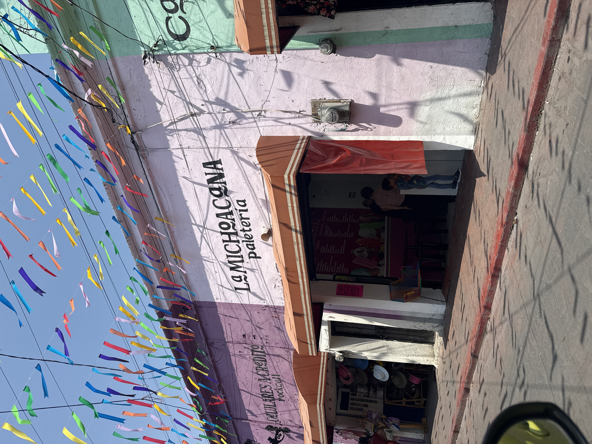 This image captures a vibrant street scene in Xochitepec, Mexico, under a clear blue sky. Numerous colorful ribbons are strung high above, stretching across the street, suggesting a festive or celebratory ambiance.
The main subject is a multi-colored building facade housing several commercial establishments. The upper portion is painted light green, followed by a pale pink section, and a purple section to the left. On the pale pink wall, the prominent text "LA MICHOACANA paleteria" is visible, identifying a well-known ice cream and popsicle shop. Its entrance is partially covered by a red tarp beneath a striped brown awning, and two people are faintly discernible inside. To the left, on the purple wall, the text "CELULARES A CREDITO rockcell" indicates a mobile phone store offering credit. Another shop with a similar striped brown awning is situated to the right of La Michoacana.
The scene is brightly lit by the sun, casting strong, distinct shadows from electrical wires, utility meters mounted on the building, and various structures onto the walls and the paved street/sidewalk. This suggests the time of day is midday or early afternoon. A red line is painted along the edge of the sidewalk or curb. In the bottom right corner, a portion of a vehicle's rearview mirror is visible. The overall impression is one of a bustling, colorful town street.