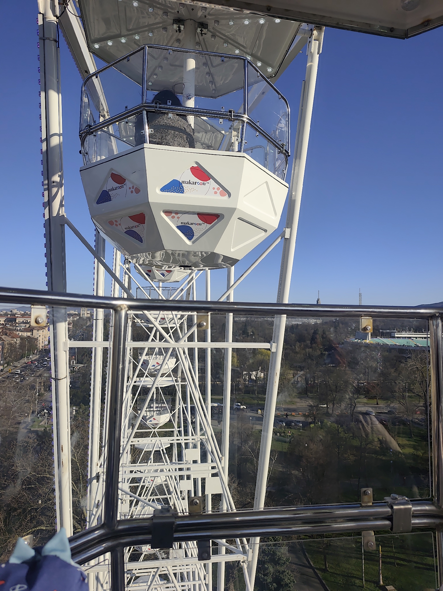 View from a Ferris wheel in Sofia, Bulgaria, showing a partial view of a passenger cabin labeled "makaron." The wheel's white metal framework is in the foreground. Beyond the wheel, the city spreads out with buildings, trees, and streets. In the distance, a stadium-like structure and towers are visible against a clear blue sky. The photo is taken through the Ferris wheel's enclosure.