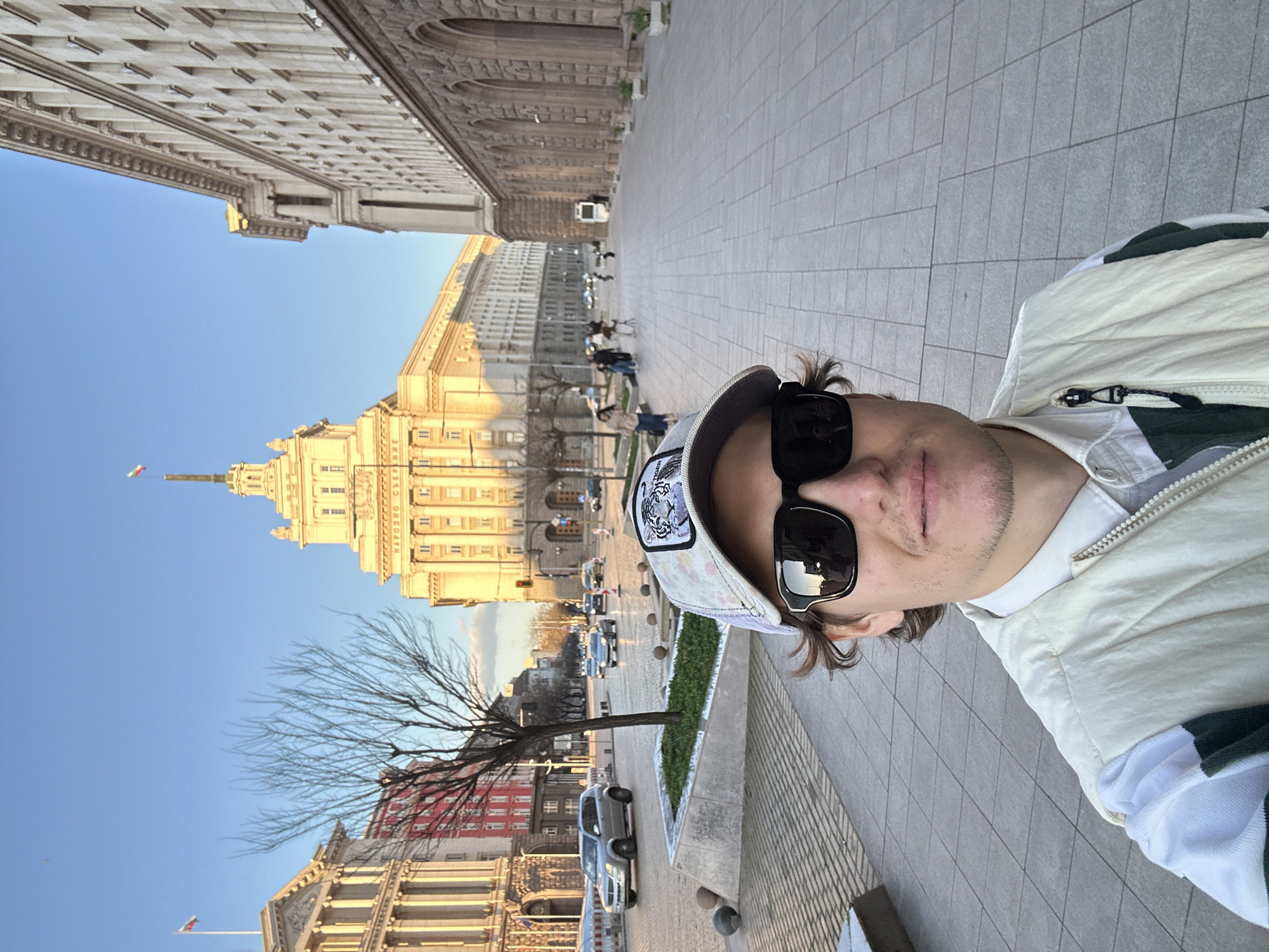A vertically oriented medium shot depicts a man in sunglasses and a baseball cap taking a selfie. The background reveals an urban scene with prominent architectural structures and vehicular traffic. Specifically, the architectural elements include detailed facades of buildings, likely governmental or public, with distinctive features such as columns, ornate windows, and spires topped with flagpoles. The buildings appear constructed from light-colored stone or concrete, illuminated by natural sunlight. The surrounding environment shows a paved walkway, green patches of manicured landscaping, and urban infrastructure such as cars and traffic signals. The overall setting suggests a location in the city of Sofia, Bulgaria, possibly near notable landmarks such as the Largo.