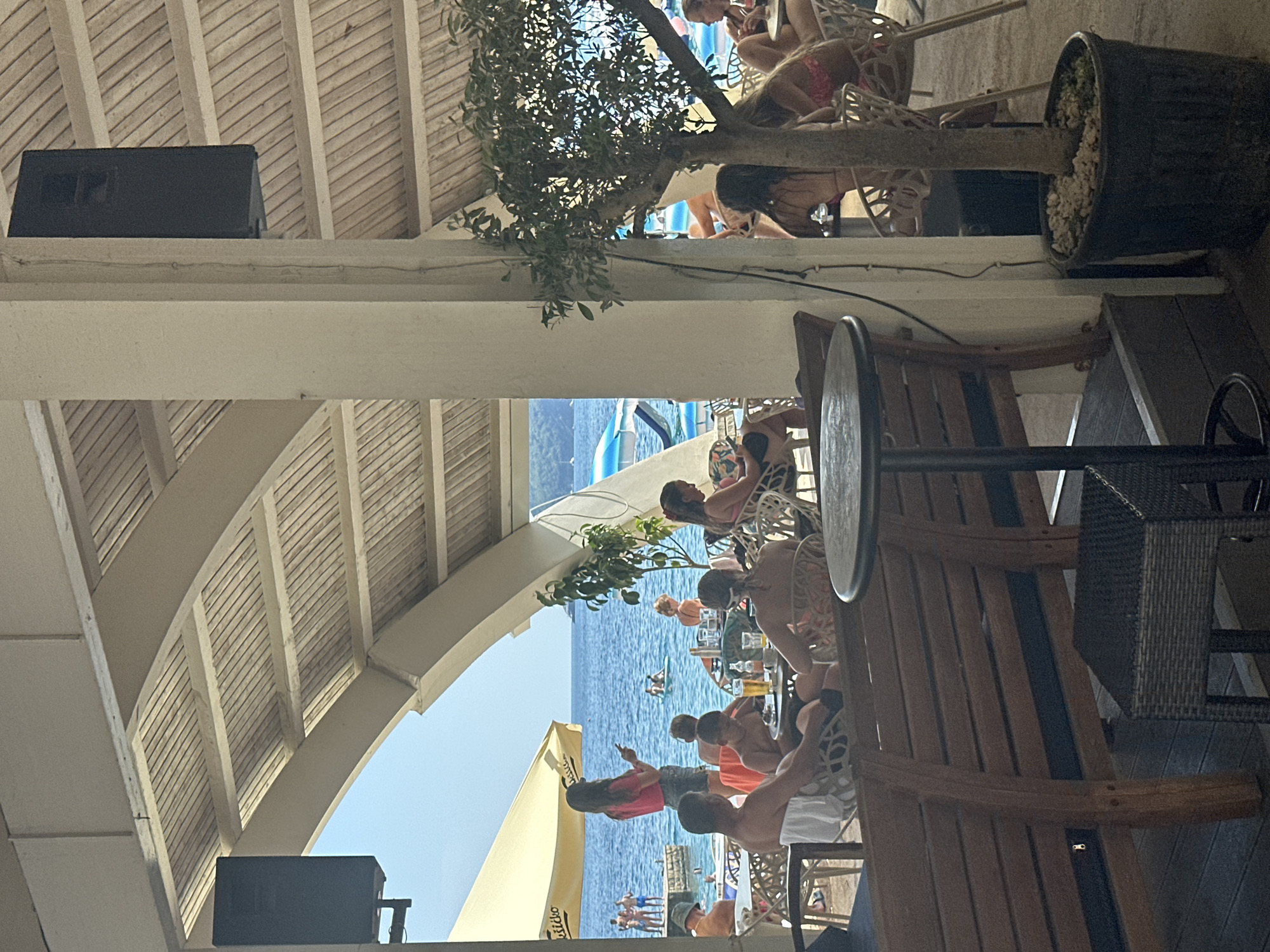 This image depicts an outdoor seating area at a beachside establishment, likely a restaurant or cafe, in Budva, Montenegro. The foreground features wooden benches and tables with chairs, some occupied by patrons. A potted tree stands near a white pillar, with a black speaker mounted on the ceiling above. In the background, people are visible on a beach and in the clear blue sea, with boats and umbrellas dotting the water. The architecture suggests a relaxed, casual atmosphere typical of a coastal vacation spot. The overall scene conveys a sense of leisure and enjoyment in a picturesque seaside environment.