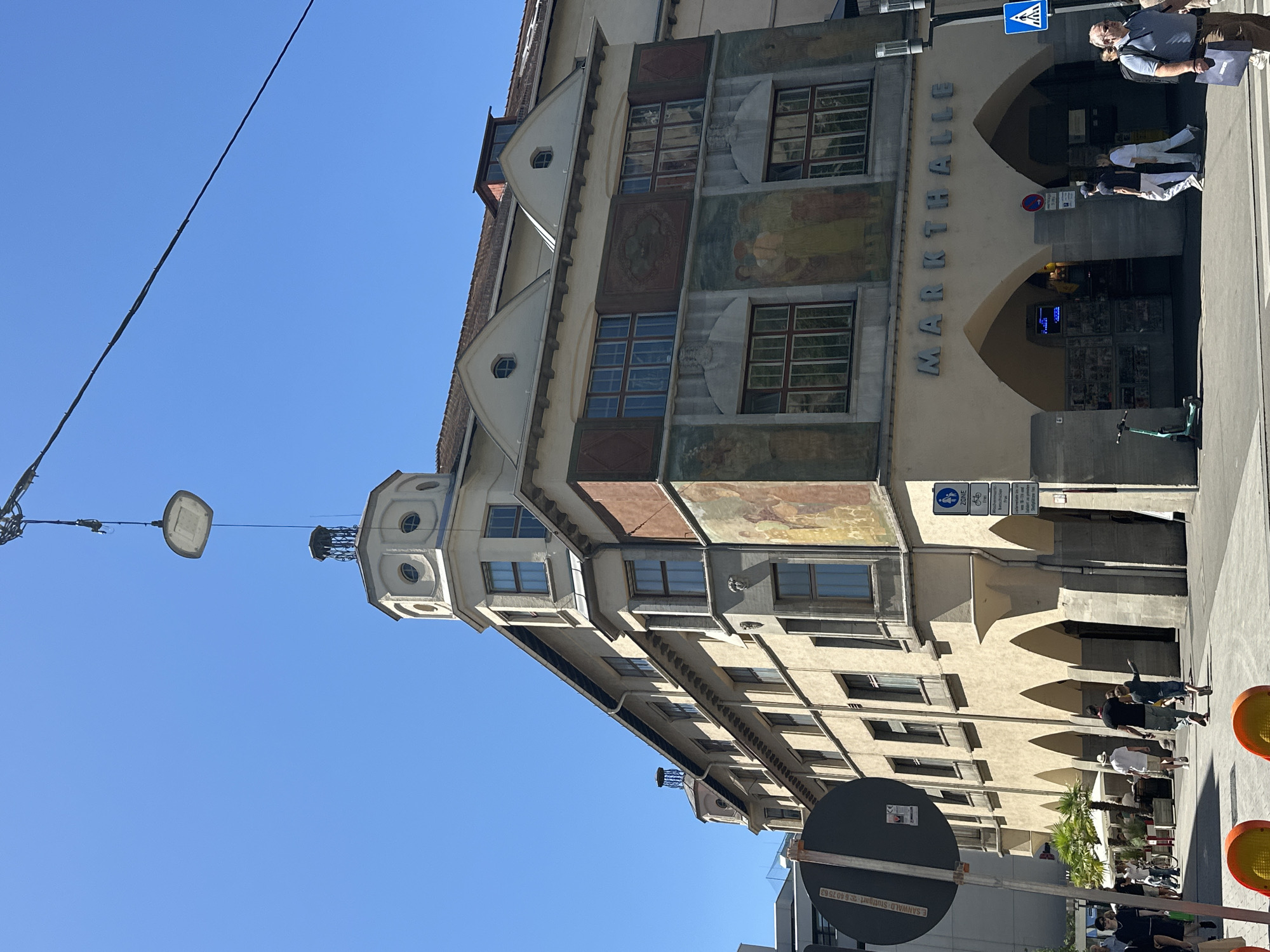 The image shows a sunny day in front of the Markthalle in Stuttgart, Germany. The Markthalle is a historic building with a distinctive architectural style, featuring arched entrances and detailed murals on its facade. A crane is visible in the sky, lowering a large, square light fixture, possibly for installation or repair. Several people are walking on the sidewalk in front of the building, some carrying shopping bags. The street is paved, and there are trees and outdoor seating areas visible, suggesting a lively urban environment. A traffic sign and a pedestrian crossing signal are also present. The sky is clear blue.