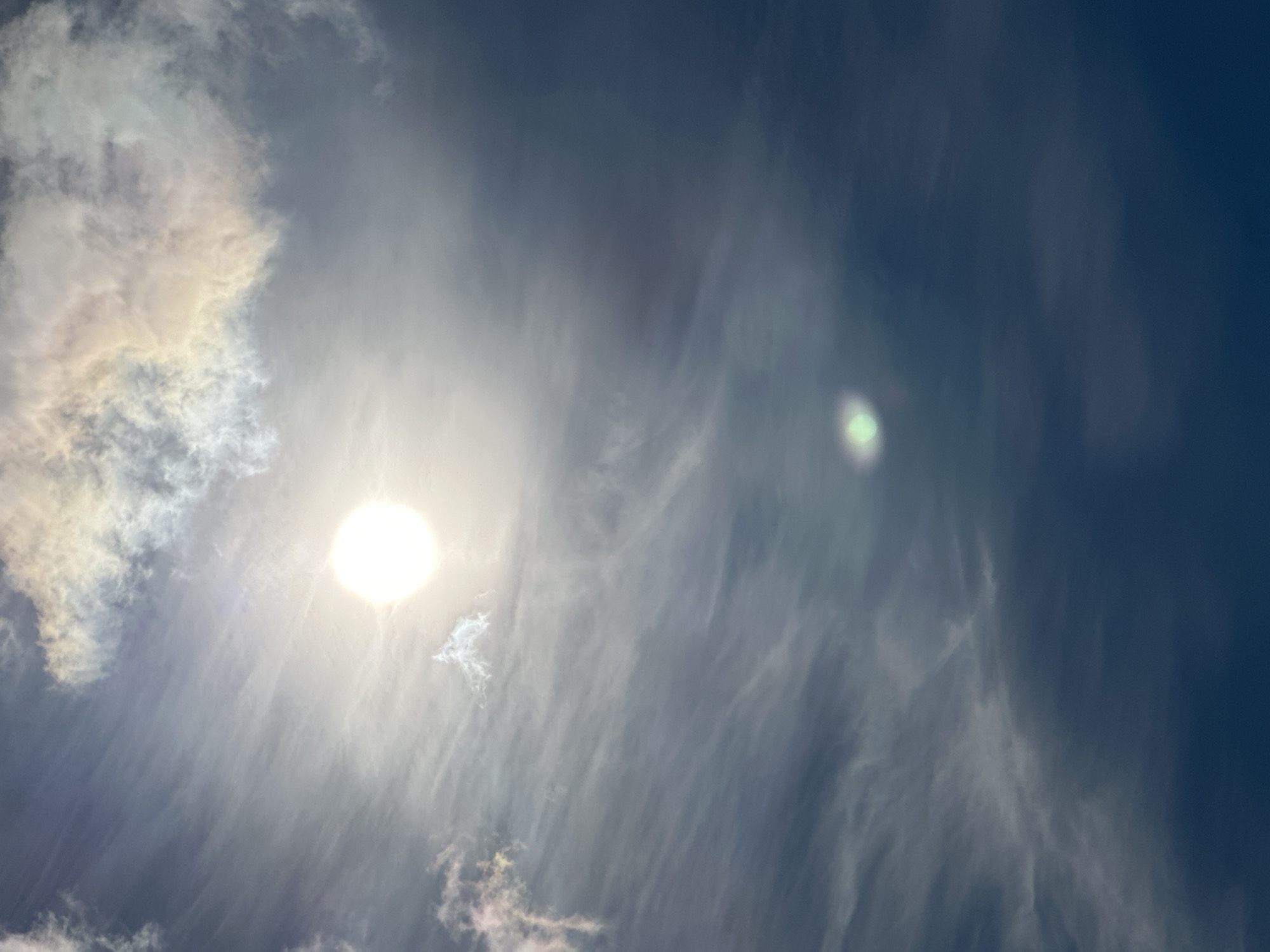 The image shows a sky with clouds and the sun. There are wispy clouds, some of which are illuminated with iridescent colors, suggesting they might be nacreous clouds. The sun is a bright, circular orb, with some lens flare visible. There is a smaller, greenish lens flare lower in the frame. The sky is a deep blue, becoming darker towards the bottom of the image. The overall impression is of a bright, possibly cold, atmospheric phenomenon. The location is identified as Podolsk, Russia.