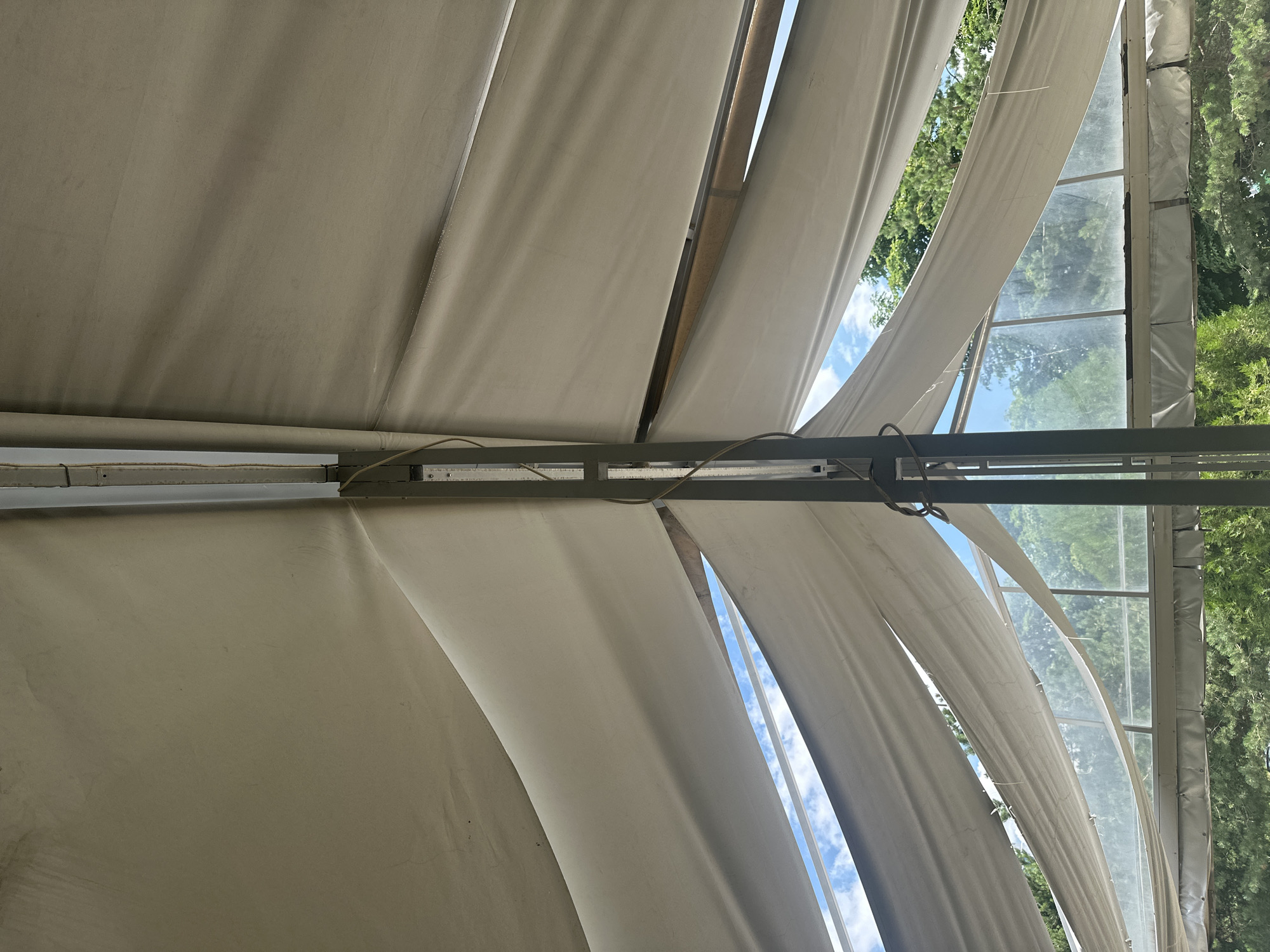 The image captures an interior view of a large tent or canopy structure, likely an event space or outdoor seating area. The focus is on the draped, off-white fabric ceiling, which is supported by a central vertical metal pole and a horizontal metal beam. The fabric is pulled taut in some areas and hangs loosely in others, creating a sense of movement and volume.
Through gaps in the fabric, a bright blue sky with scattered white clouds is visible, suggesting an outdoor setting on a clear day. Beyond the tent structure, a backdrop of lush green trees and foliage is seen, indicating a location surrounded by nature. The lower portion of the image reveals a transparent or translucent wall, possibly made of glass or plastic, which allows a view of the surrounding trees. There are also some metallic frames and wires integrated into the tent's support system.
The scene is devoid of any people. The overall impression is one of an open, airy space designed for outdoor gatherings, offering shelter while maintaining a connection with the natural environment. The specific location in Sofia, Bulgaria, is not discernible from the visual information provided.