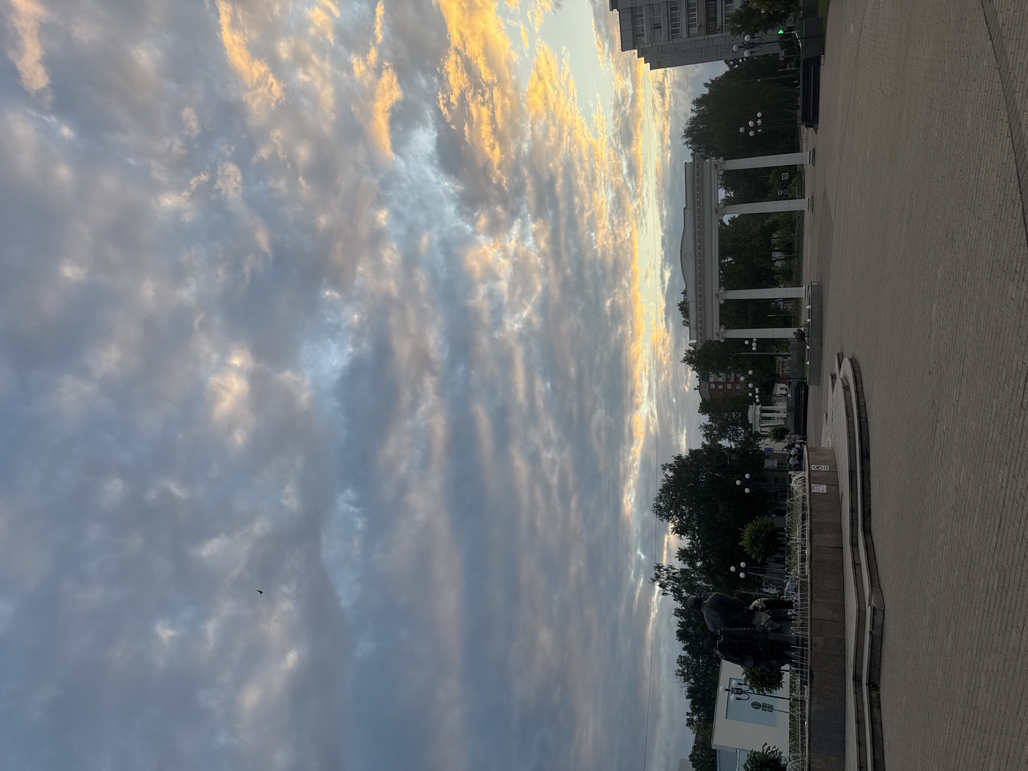 The image depicts a public square in Chaikovsky, Russia, at dusk. The sky is filled with dramatic clouds, tinged with hues of orange and yellow from the setting sun. In the foreground, a paved plaza leads to a large, multi-tiered fountain with a dark statue at its center, possibly depicting a historical or allegorical scene. Several jets of water are visible in the fountain. Beyond the fountain, trees line the square, and a neoclassical archway with white columns stands prominently. To the right, a modern high-rise building contrasts with the more traditional architecture. A few scattered people are visible in the background, suggesting a leisurely evening in the city. The overall scene conveys a sense of calm and twilight beauty.