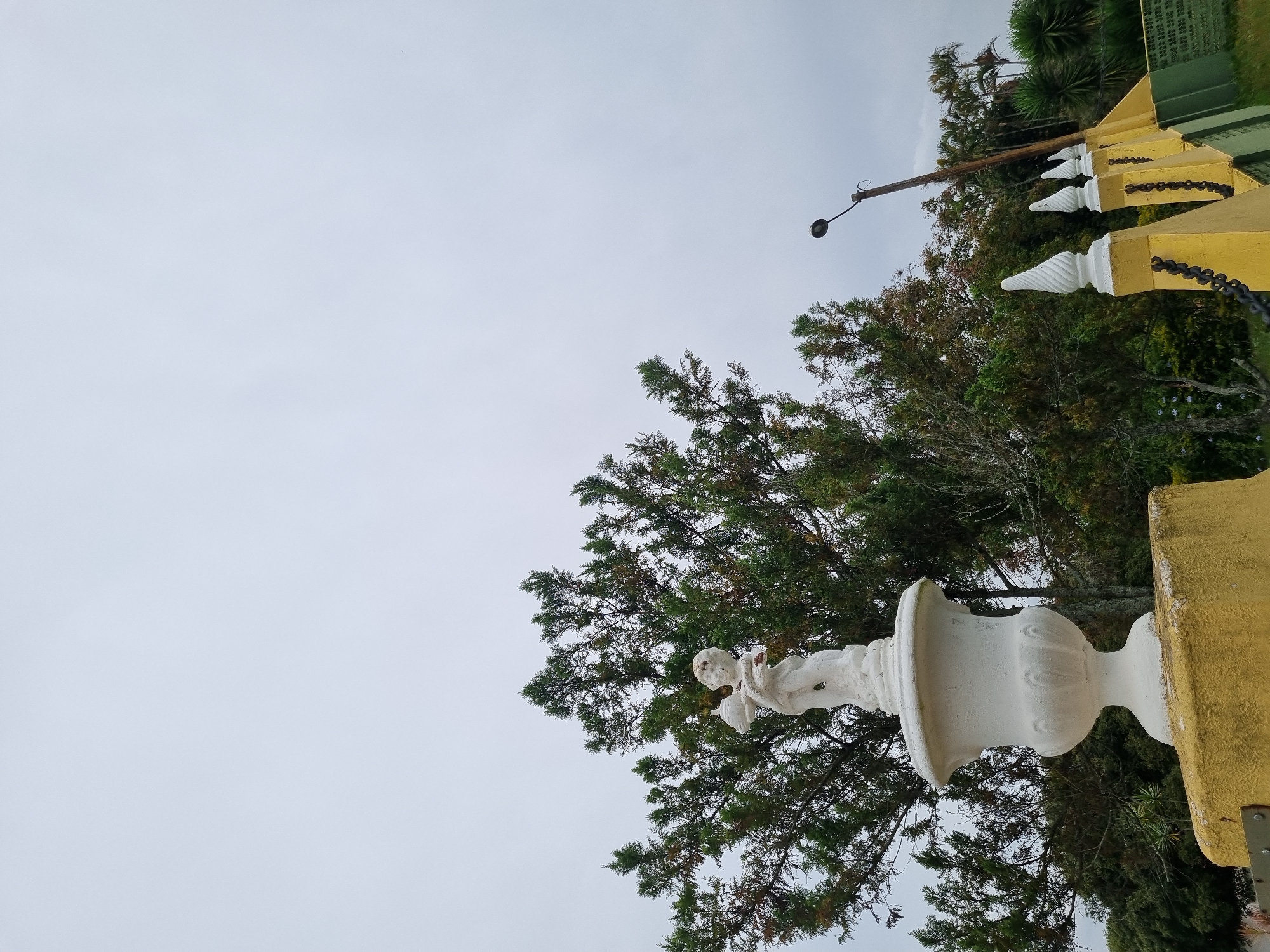This image captures an overcast day in Laranjal Paulista, Brazil, focusing on an outdoor decorative arrangement. A white classical urn, topped with a white statue of a cherub holding a spherical object, stands prominently on a textured yellow pedestal. Behind this structure, a large evergreen tree with dense foliage dominates the left side of the frame. To the right, a slender lamppost with a dark cap extends upwards against the pale sky. Further to the right, a row of yellow pillars, adorned with white conical finials and black chains, creates a border. Beyond these elements, a dense collection of palm trees and other greenery suggests a park or garden setting. The overall impression is one of a tranquil, possibly historical, outdoor space.