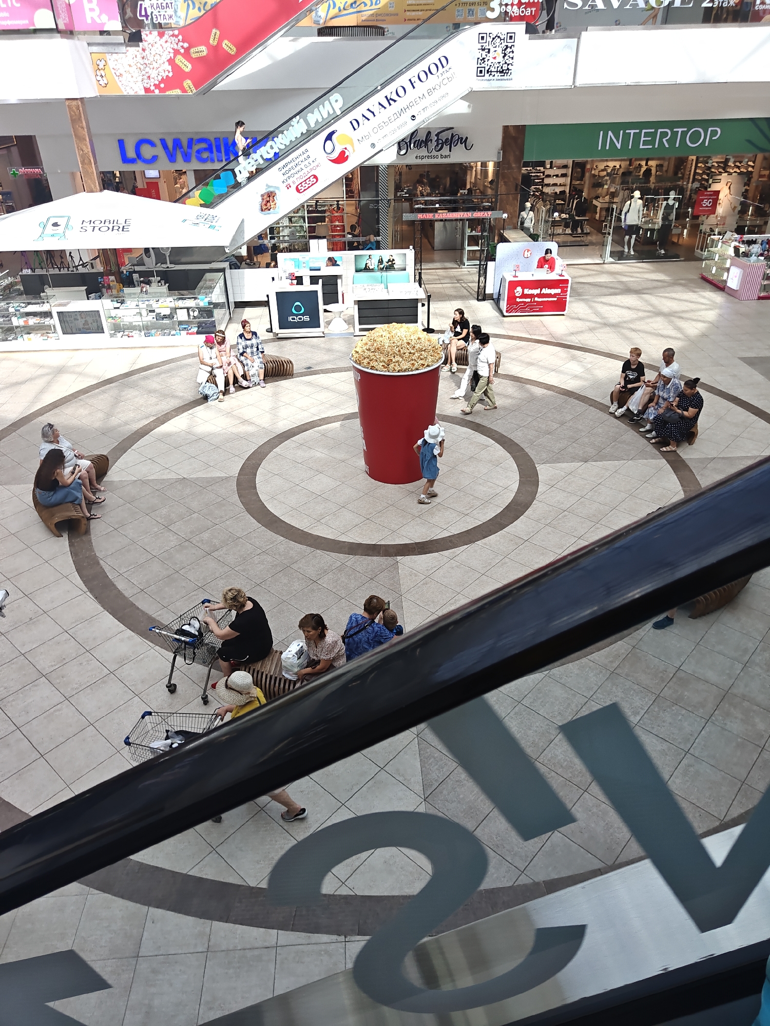 This is an aerial, high-angle shot taken inside a shopping mall. In the foreground, a black railing or barrier obscures the bottom portion of the image, casting a shadow across the tiled floor.
In the mid-ground, a large red cup filled with popcorn is a central focal point. A young girl in a blue dress and white hat stands near the popcorn cup, looking towards it. Several groups of people are seated on curved benches arranged in concentric circles on the patterned tile floor.
Further back, escalators are visible, with signage for "LC Waikiki" and "DAYAKO FOOD" on one. Shops such as "INTERTOP" are also visible. People are seen walking and sitting in various areas of the mall. The overall environment appears to be a bustling indoor shopping center. The image is confirmed to be from Petropavl, Kazakhstan.