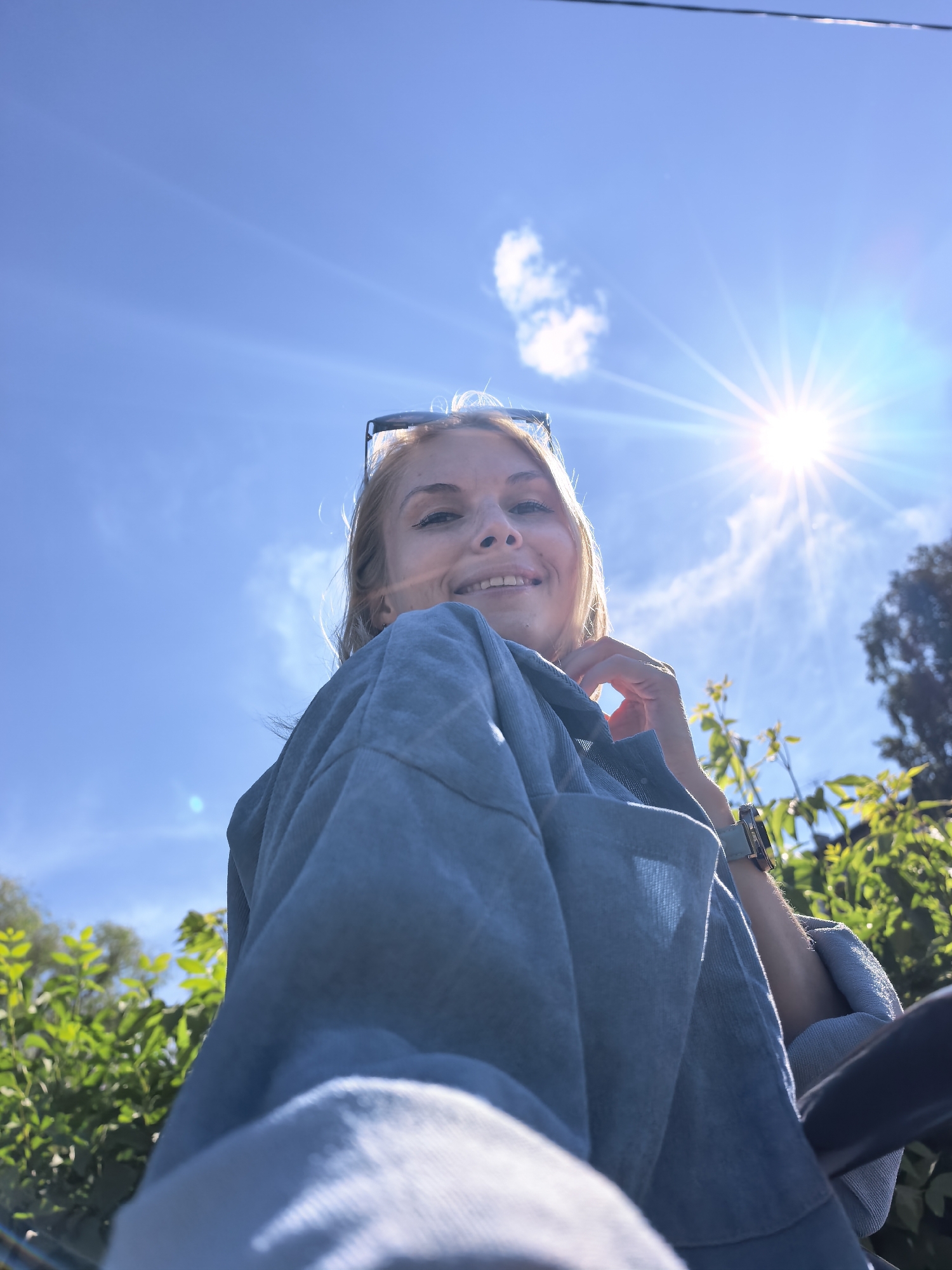 A low-angle outdoor portrait captures a Caucasian woman smiling directly towards the camera. She has blonde hair and wears sunglasses on top of her head. Her left wrist displays a watch with a square face and a light-colored band. She is dressed in a light blue, collared garment. The background features a clear blue sky with a prominent sunburst and lens flares in the upper right, and a few white wispy clouds. Lush green foliage and the dark silhouette of a tree are visible in the lower and right portions of the frame. The scene is located in Sterlitamak, Russia.