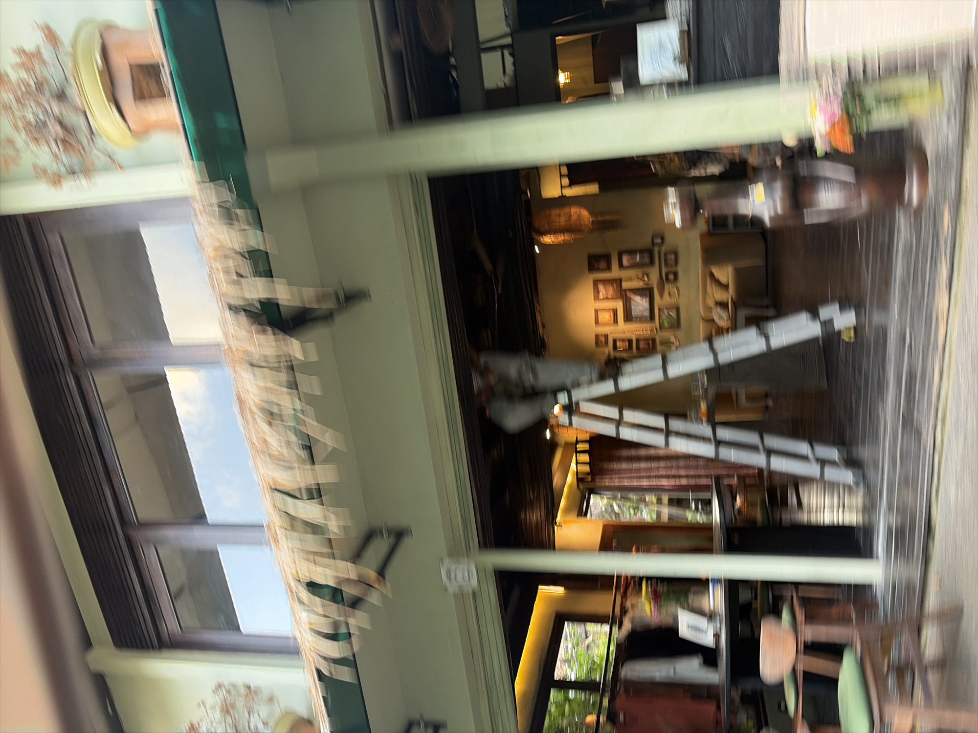 The image depicts the exterior of a shop or establishment in Ubud District, Indonesia. A person, wearing a gray uniform, is on a tall, aluminum ladder, appearing to be performing some maintenance or installation work on the facade of the building. The building has a light green exterior wall and large, dark-framed windows. A thatched awning or decorative element hangs above the entrance, with strips of material extending downwards. Inside, the shop appears to be decorated with numerous framed pictures on the wall. The entrance is open, revealing shelves with items, clothing racks with apparel, and furniture. A glass vase with flowers is visible near the entrance. The overall impression is of a retail space in a tropical location.