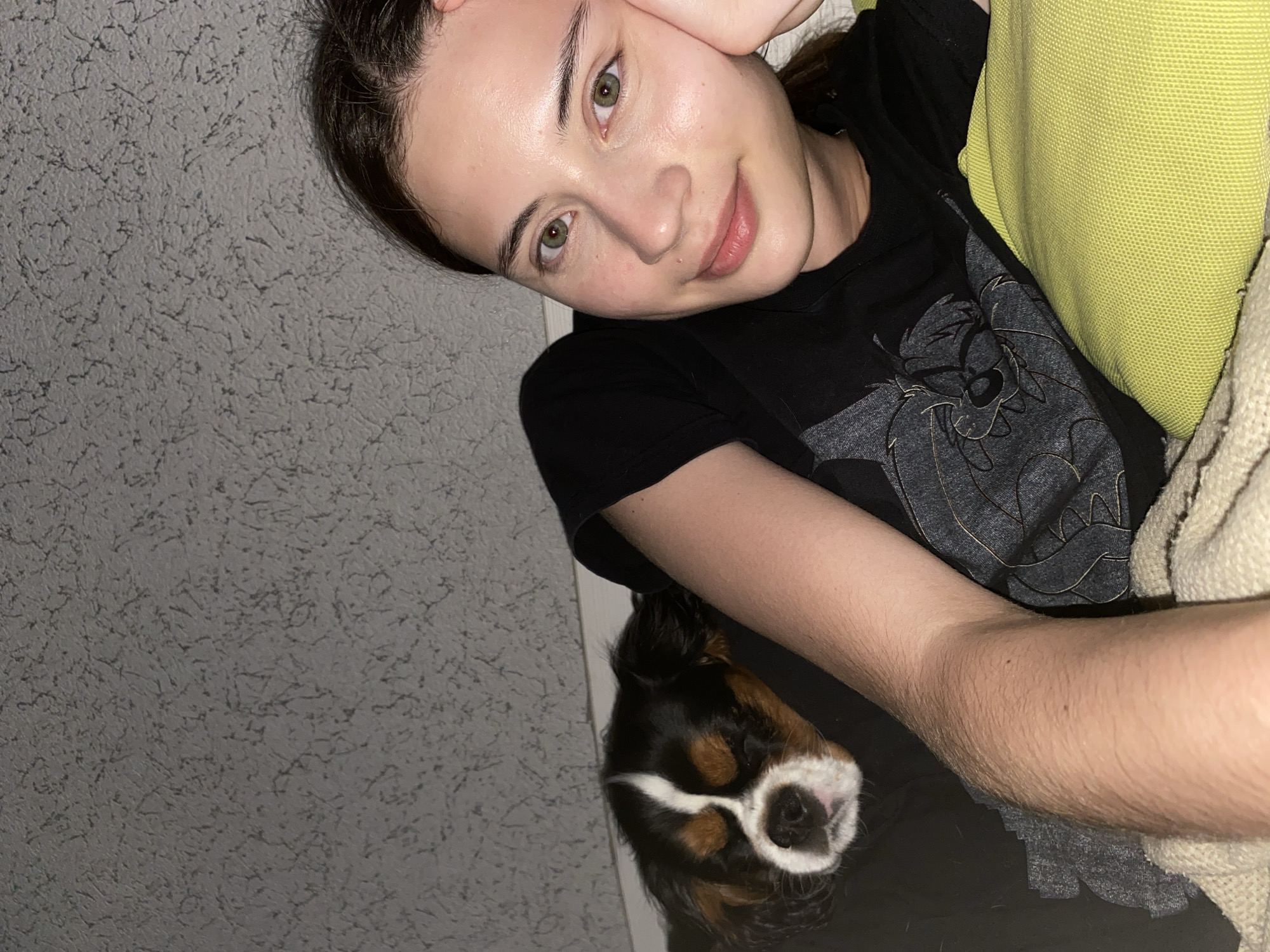 A young woman with brown hair and green eyes is lying down next to a sleeping tri-color Cavalier King Charles Spaniel. She is wearing a black t-shirt with a graphic design. Her left arm is resting on a green cushion, and her right hand is propping up her head. The dog is nestled against her left side. The background appears to be a textured grey wall. The scene is indoors, and the lighting suggests it is nighttime. The setting is not identifiable as being in Votkinsk, Russia.