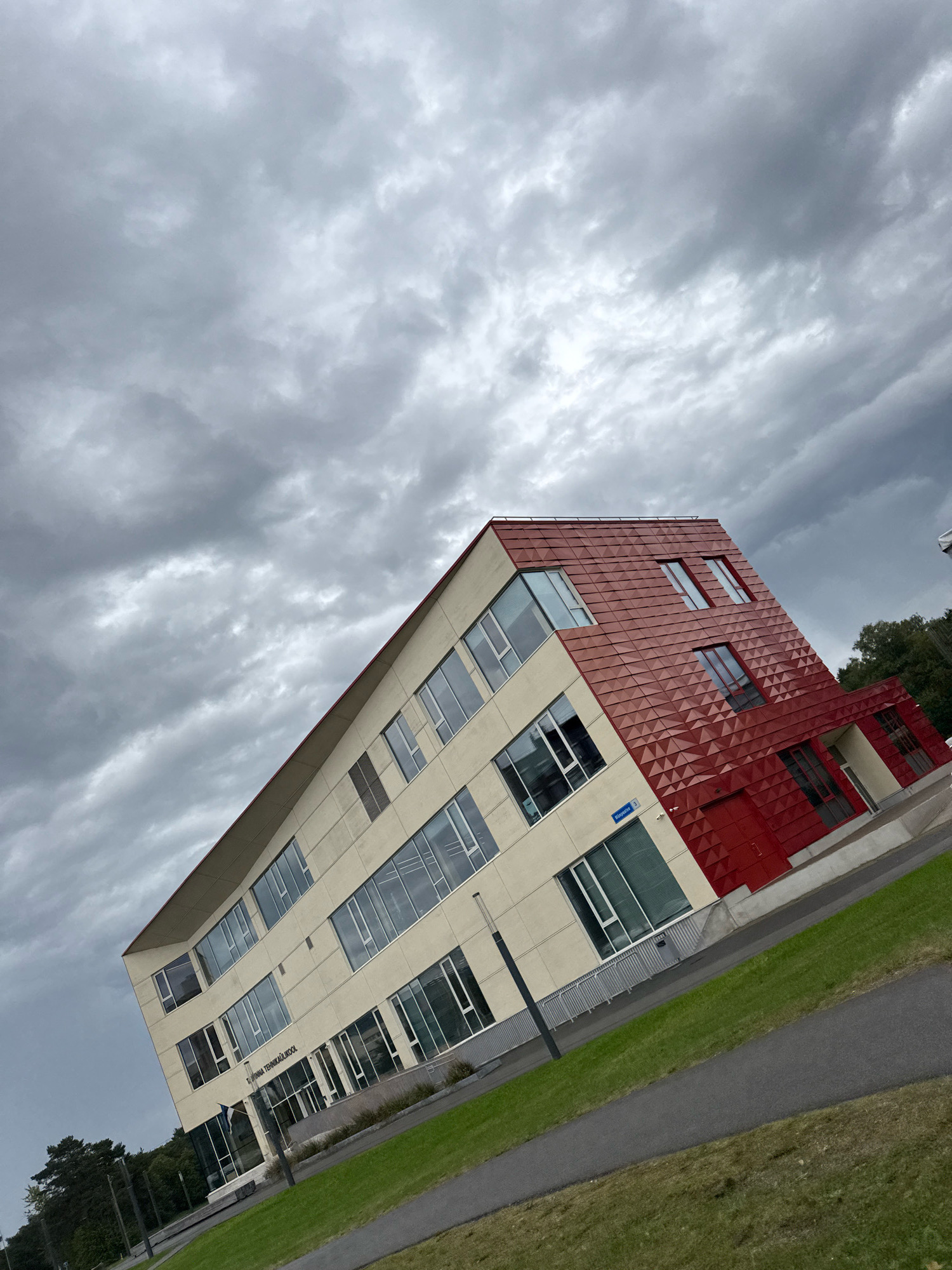 The image shows a modern, multi-story building with a cream-colored facade and a distinctive red, geometric-patterned section. The building has numerous large windows. A street sign reads "Päikese" and "5". The building is situated in an area with green grass and a paved pathway or road. The sky is overcast with dark, heavy clouds. The building is identified as "Tallinna Tehnikaülikool" which translates to Tallinn University of Technology, indicating its location in Tallinn, Estonia. The image is taken from a slightly elevated perspective, looking up at the building.