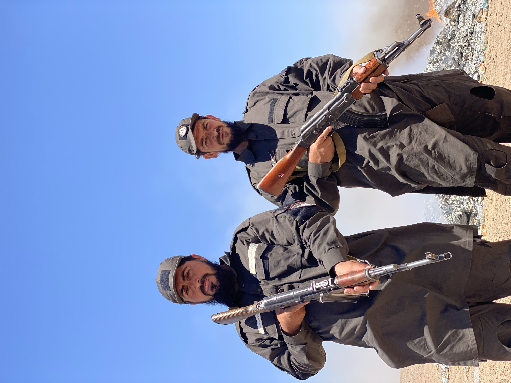 Two men in dark uniforms and caps stand outdoors under a clear blue sky, each holding an AK-47 rifle. They are in a dusty, outdoor environment with a large pile of debris, some of which appears to be burning, in the background. The man on the left has a full beard and is looking directly at the camera. The man on the right has a shorter beard and is also looking at the camera. Both men wear dark, long-sleeved shirts with patches on the sleeves and chest, and dark pants. The ground is a light brown, dry earth.