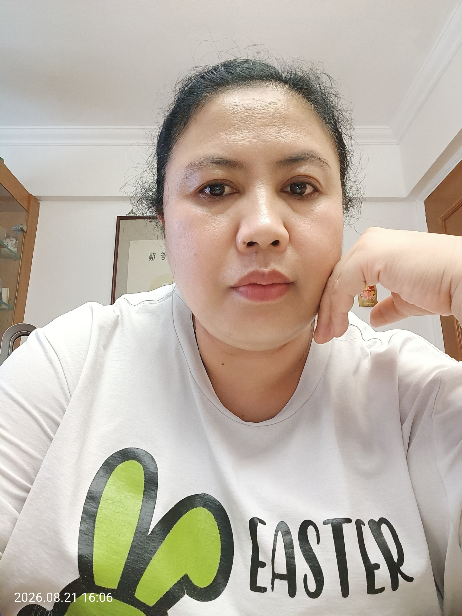 A close-up, head-and-shoulders selfie of an East Asian woman is presented. She is looking directly at the camera with her right hand supporting her chin. She is wearing a white t-shirt with a graphic design featuring green bunny ears and the word "EASTER" printed in black. The background shows a white wall with molding near the ceiling, a framed picture on the wall, and a portion of a wooden cabinet. The timestamp "2026.08.21 16:06" is visible in the lower-left corner of the image. There is no discernible indication of the location within Hong Kong, China.