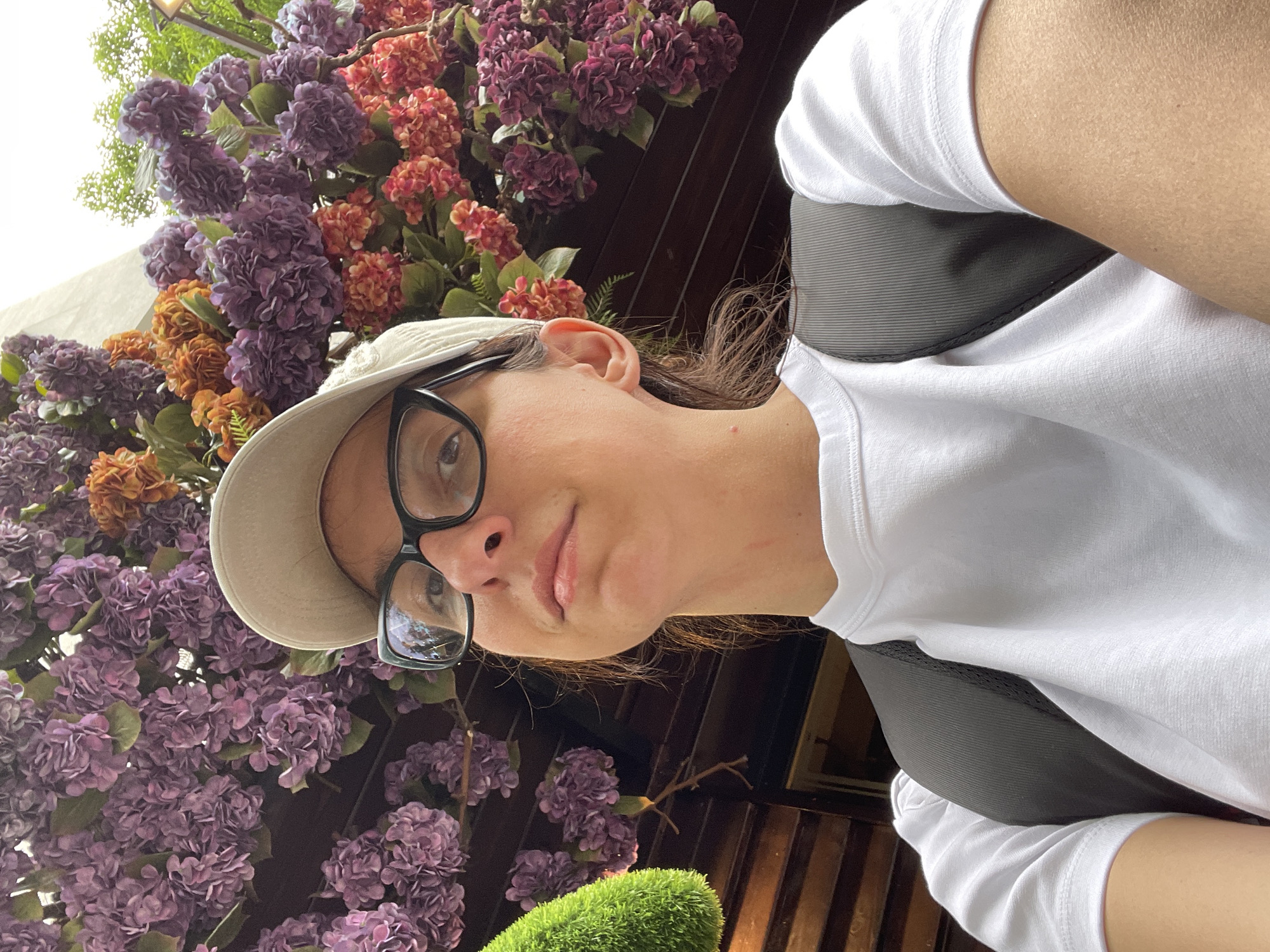 A person wearing a white T-shirt and a light-colored baseball cap poses for a selfie. The person is also wearing glasses. Behind the person is a wall of flowers in shades of purple, orange, and pink. The wall appears to be part of an outdoor setting, possibly in the city of Sofia, Bulgaria. The exact location within Sofia cannot be determined from the image. A piece of green vegetation is visible at the bottom of the frame.