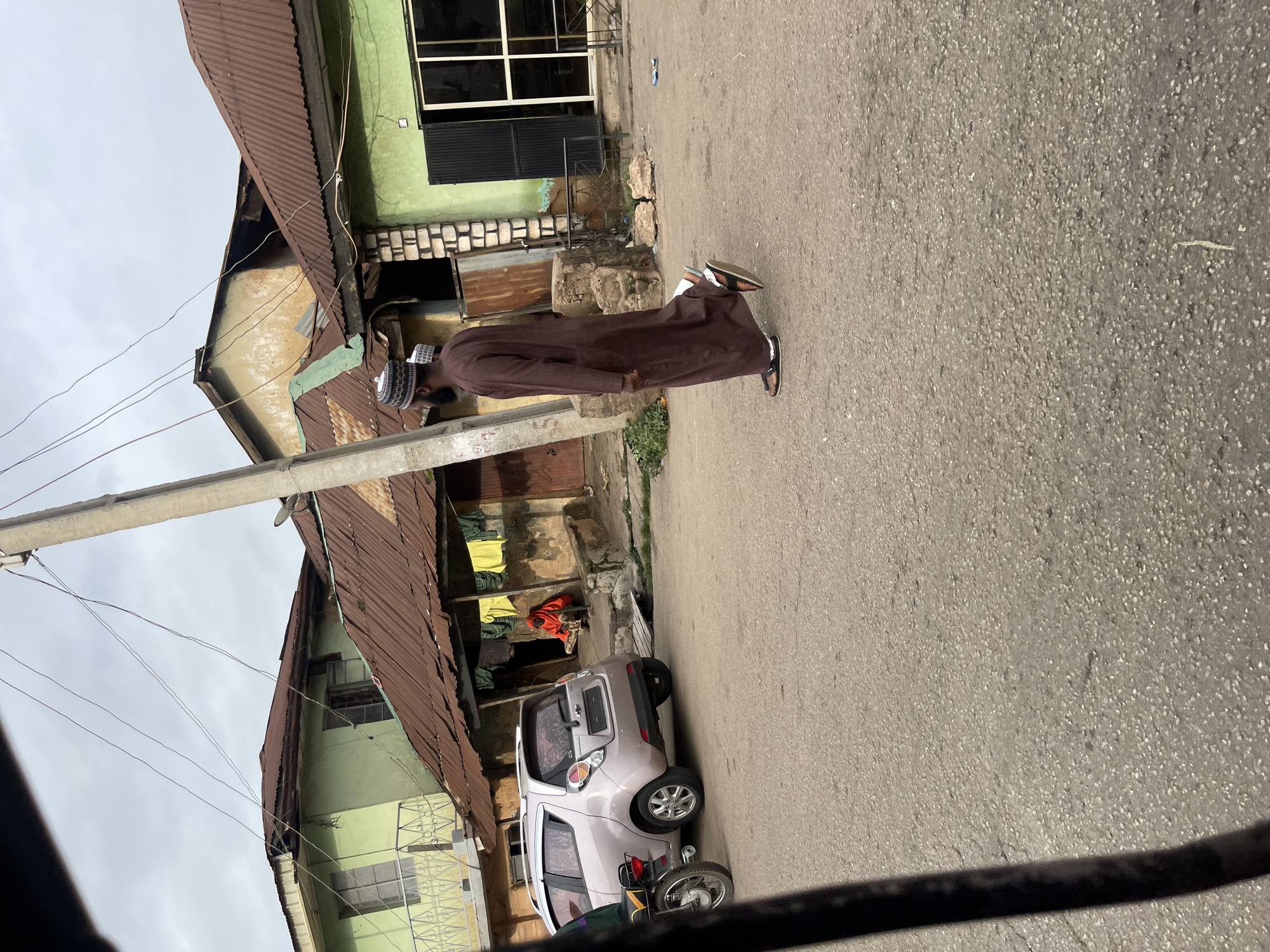 A man in a brown robe and a patterned cap walks on a paved street in front of buildings. He is moving from left to right in the image. To his left is a light pink hatchback car and a motorcycle. Behind the car, a person in an orange garment sits in an open doorway of a building. The buildings have corrugated metal roofs and some have green facades. Power lines are visible overhead, strung between a concrete pole and other poles out of frame. The scene appears to be in an urban environment with a cloudy sky.