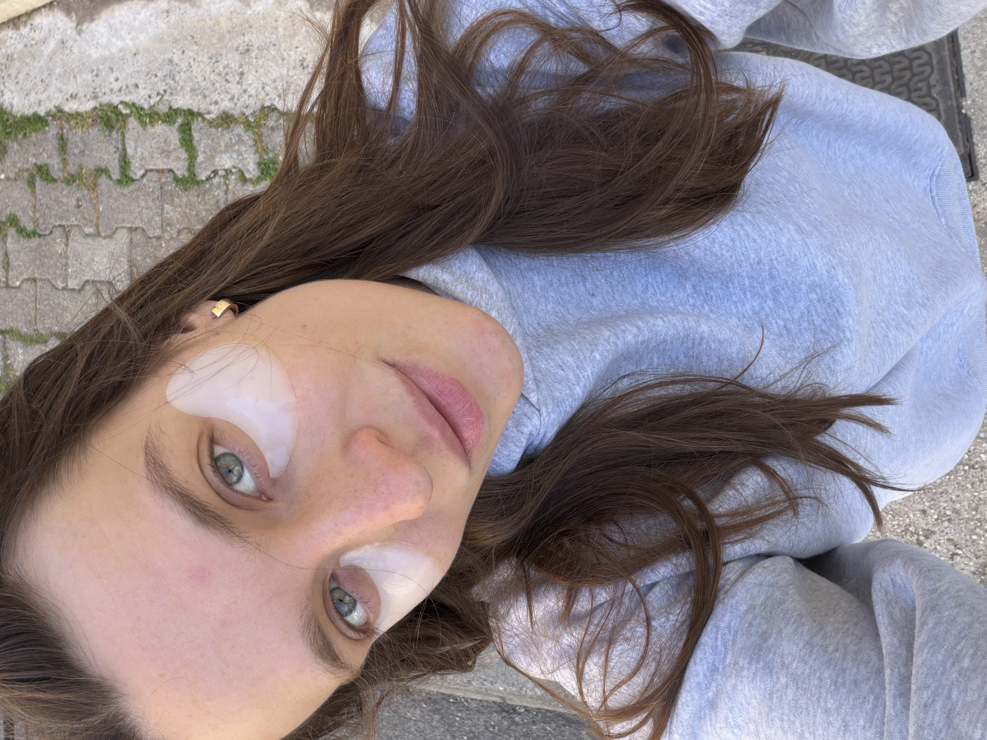 The image displays a close-up, overhead view of a young person, likely female, lying outdoors. The subject has long, wavy brown hair spread around their head and shoulders. They are wearing a light grey hooded sweatshirt.
Their face is angled upwards towards the camera, revealing light blue or green eyes and a subtle, neutral expression with slightly parted lips. Two white, translucent under-eye patches are prominently placed beneath each eye, suggesting a skincare routine. A small, gold-colored hoop earring is visible on their left ear. The skin shows a few freckles.
The background consists of light grey paving stones with patches of green grass growing in between, indicating an outdoor setting such as a pathway or patio, possibly in Budva, Montenegro. The lighting is natural and bright, consistent with daytime. The person appears relaxed, engaging in a moment of self-care or leisure. No text is visible in the image.